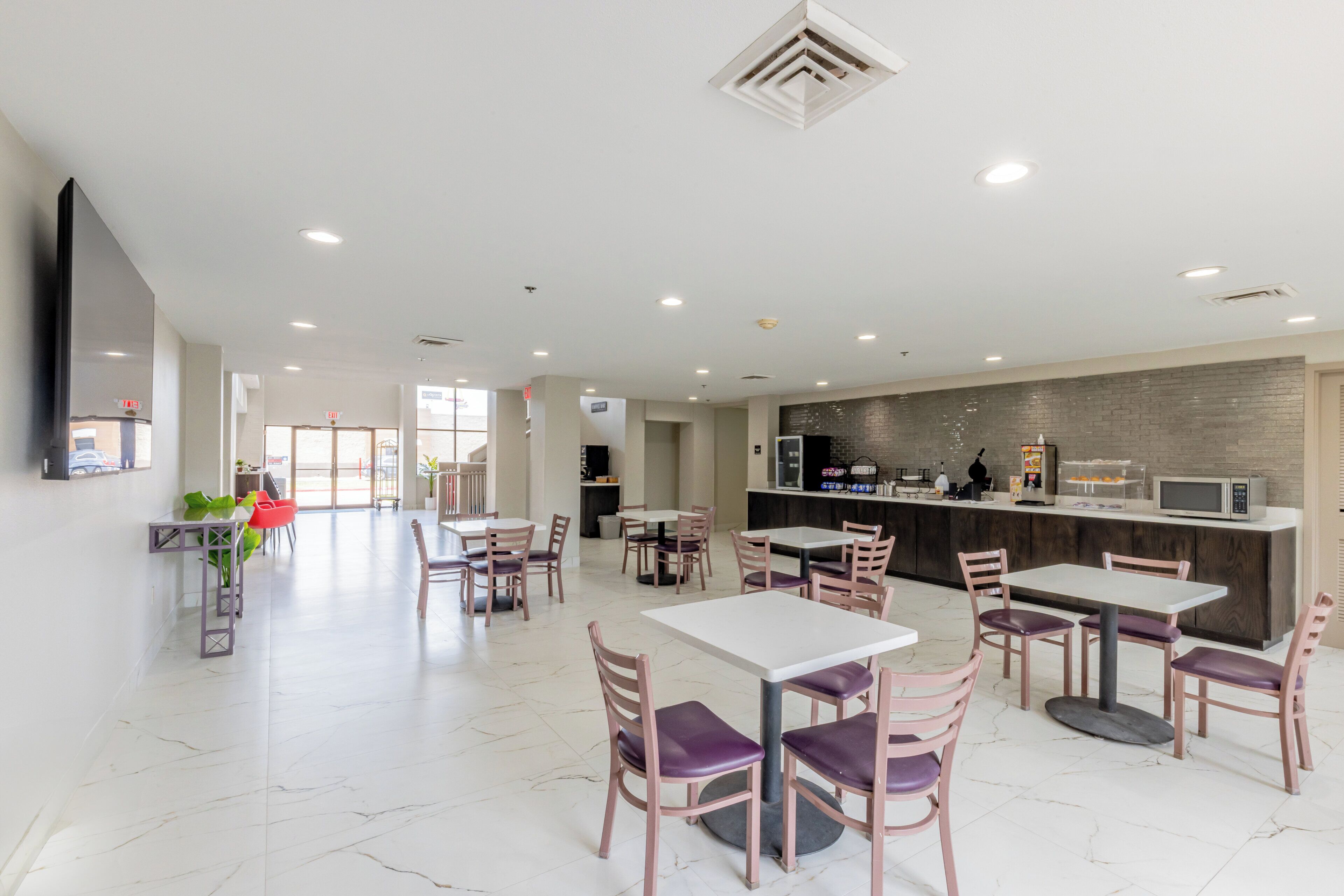 Un espace repas moderne avec des tables et des chaises blanches, un coin cuisine doté d’un mur de fond en briques et une grande fenêtre laissant entrer la lumière naturelle.