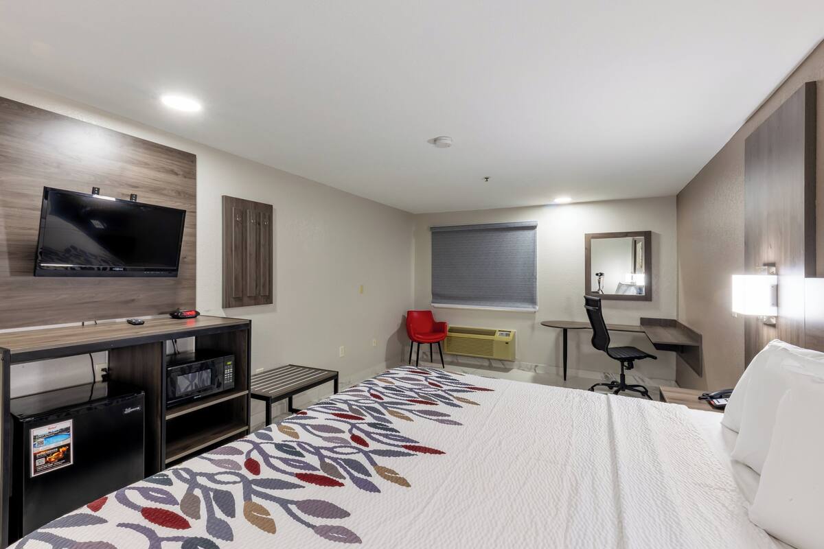 A hotel room with a bed, a desk with a computer, a TV, a microwave, and a red chair.