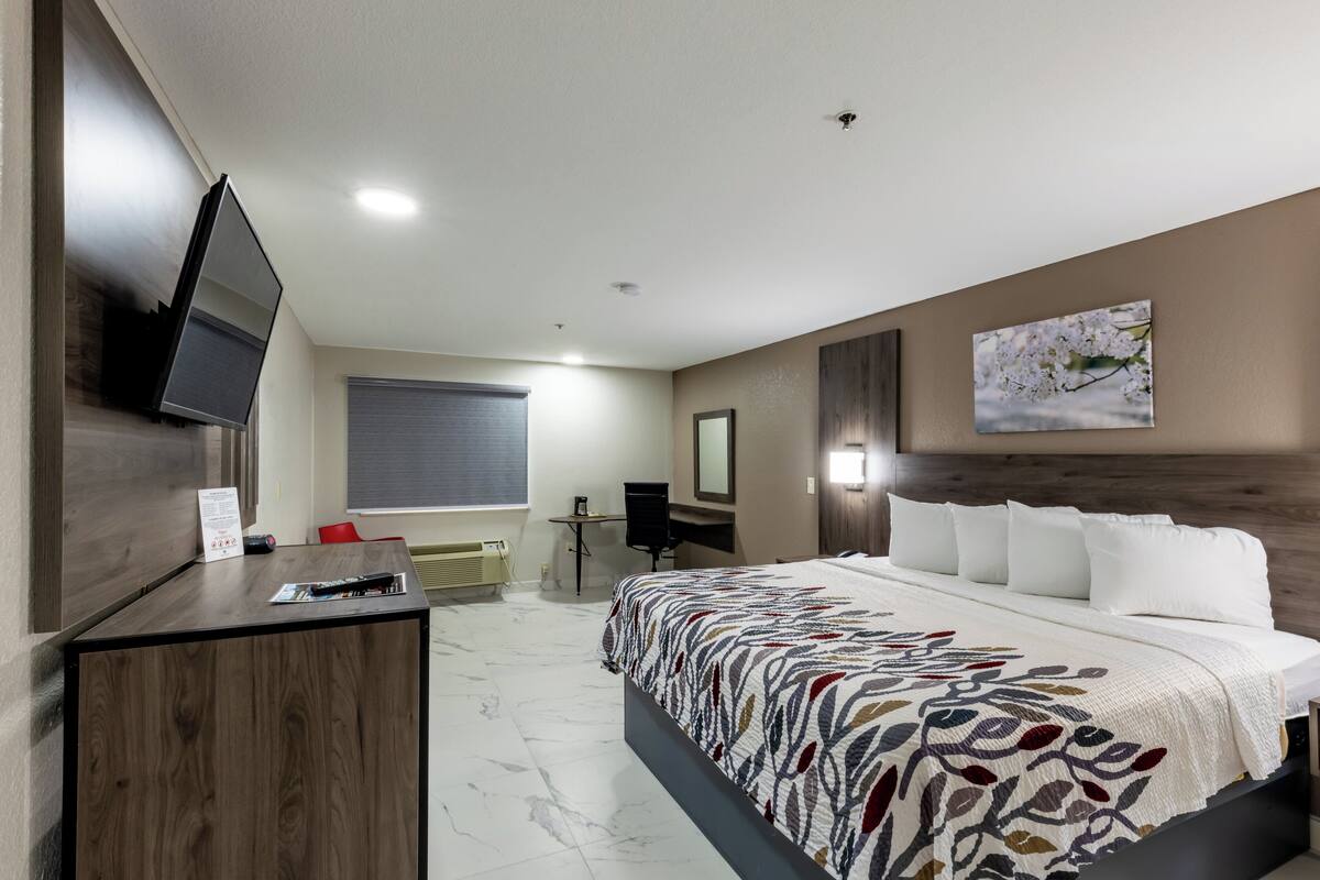 Una habitación de hotel con cama, escritorio, silla, televisión y ventana con persianas.