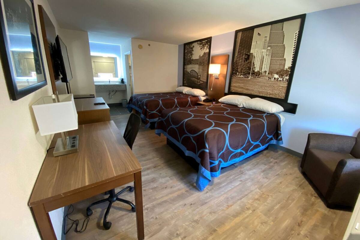 Ein Hotelzimmer mit zwei Betten, einem Schreibtisch, einem Stuhl, einem Fernseher und einem Fenster.
