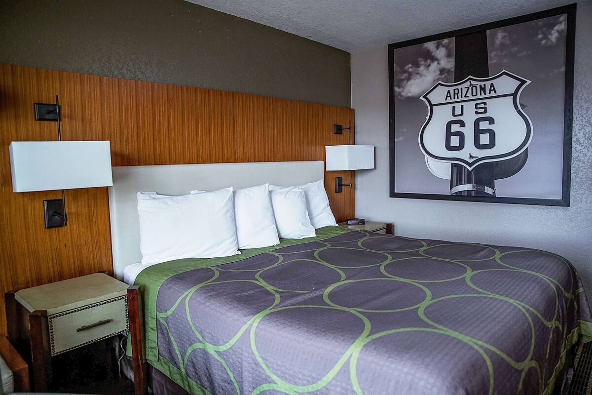 A hotel room with a wooden headboard, a bed with a purple and green patterned bedspread, two bedside lamps, and a wall-mounted sign of Arizona US 66.