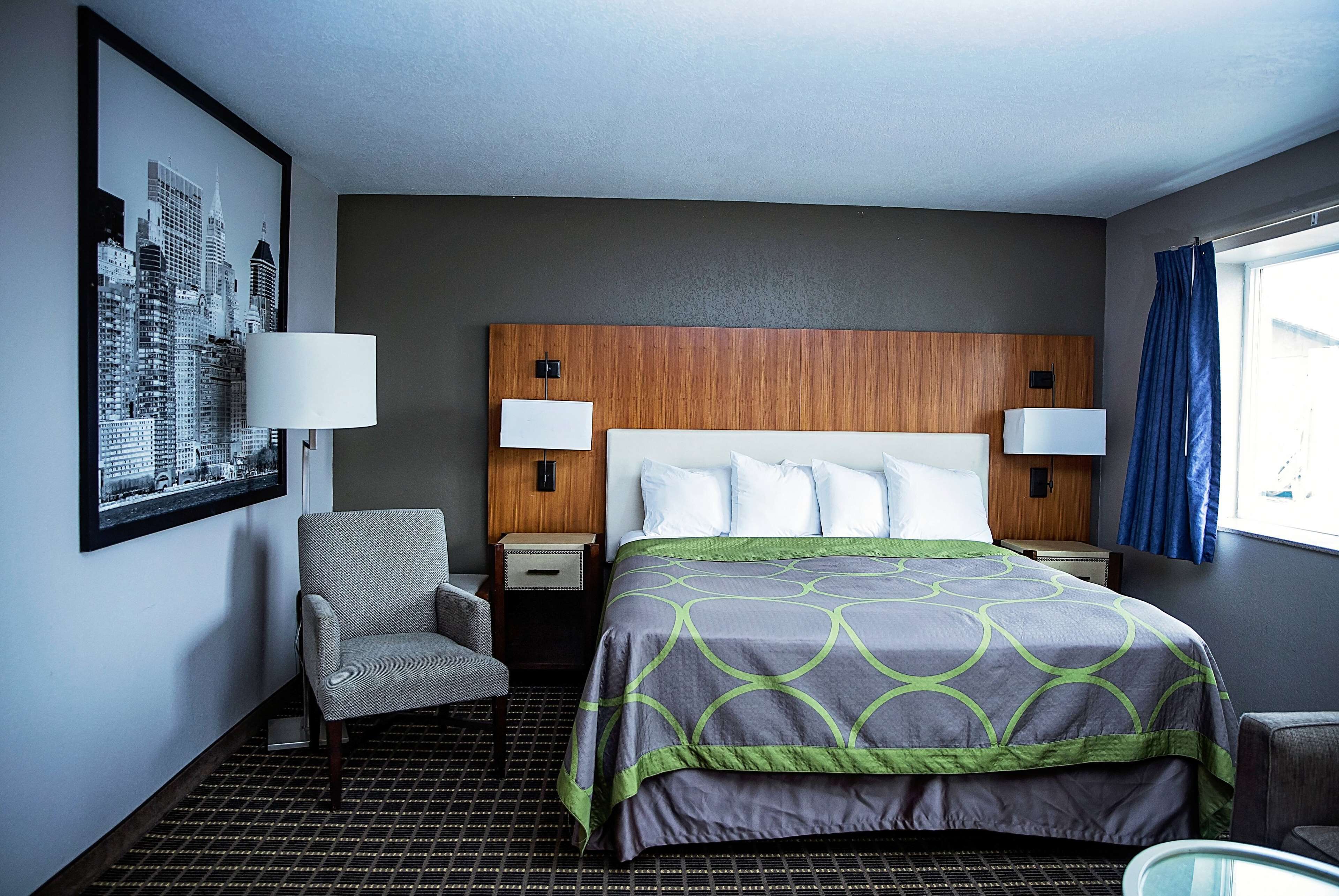 Habitación de hotel con una cama grande, dos mesitas de noche, una silla, una obra de arte con paisaje urbano en la pared y una ventana con cortinas.