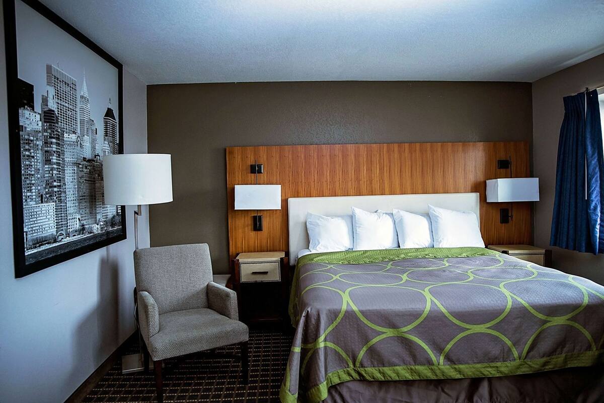 A hotel room with a bed, two bedside lamps, a chair, a nightstand, a framed cityscape picture, and a window with curtains.