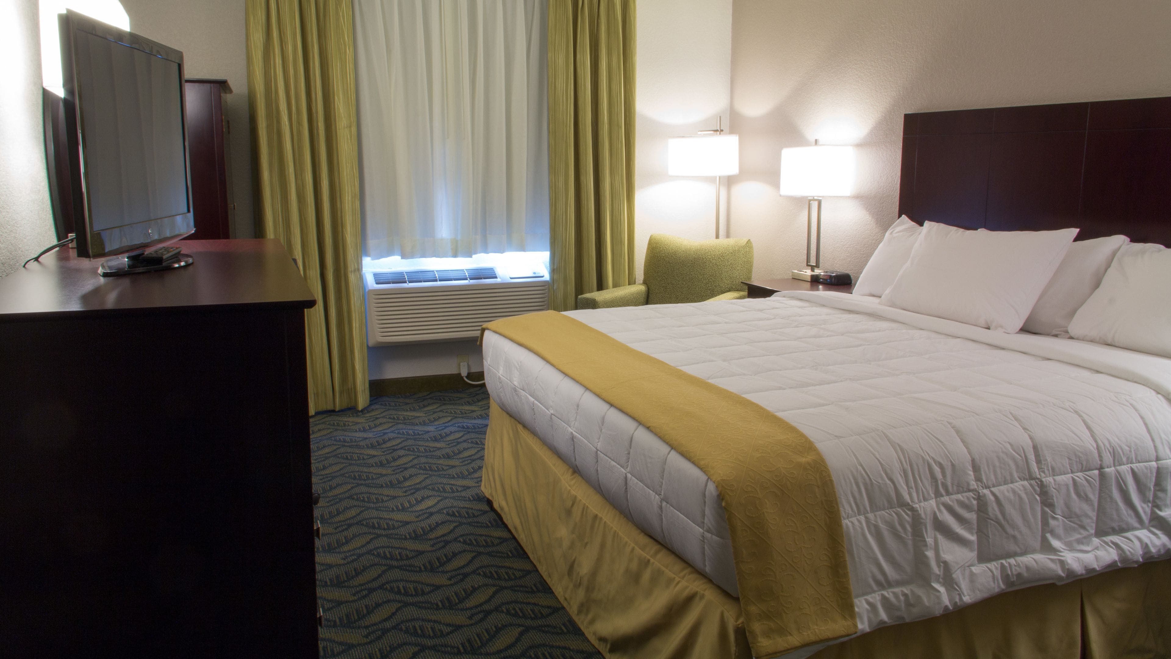 A hotel room with a bed, a desk with a television, a chair, two bedside lamps, and a window with curtains.