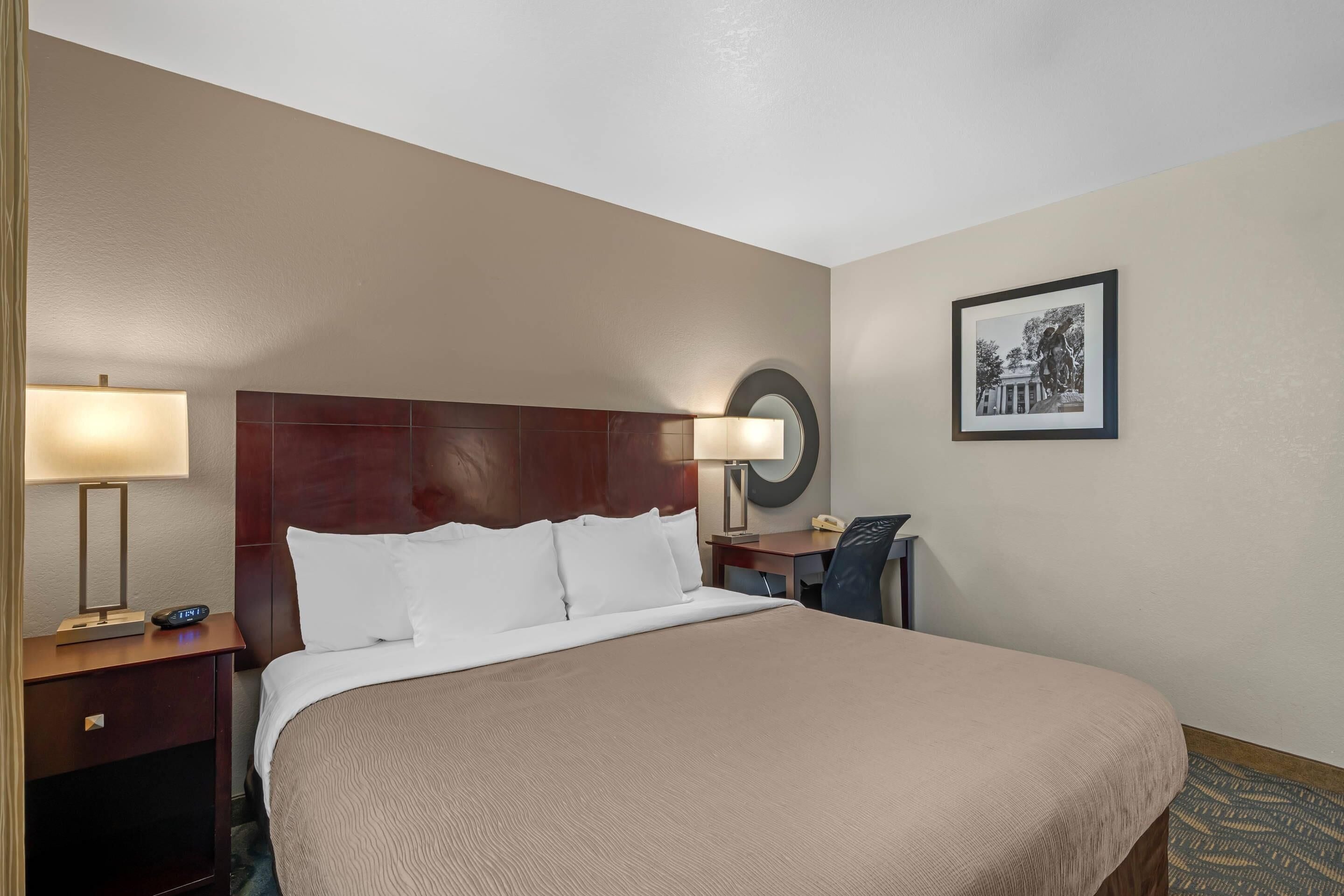 A hotel room with a large bed, a wooden headboard, a desk with a lamp, a telephone, and a framed picture on the wall.