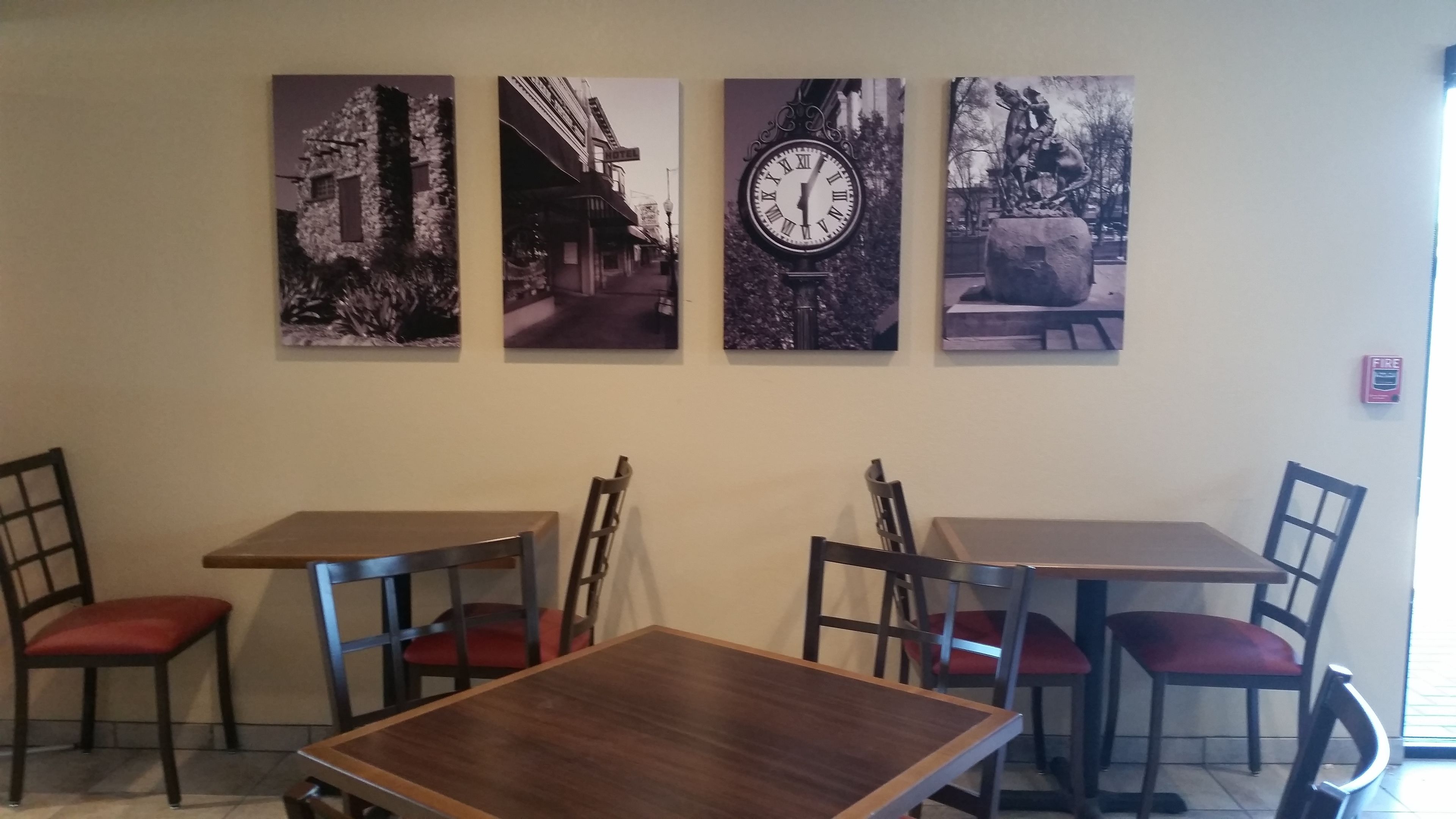 Un espace repas comprenant quatre tables et des chaises, chacune agrémentée de coussins rouges. Quatre tableaux encadrés sont accrochés au mur, au-dessus des tables.
