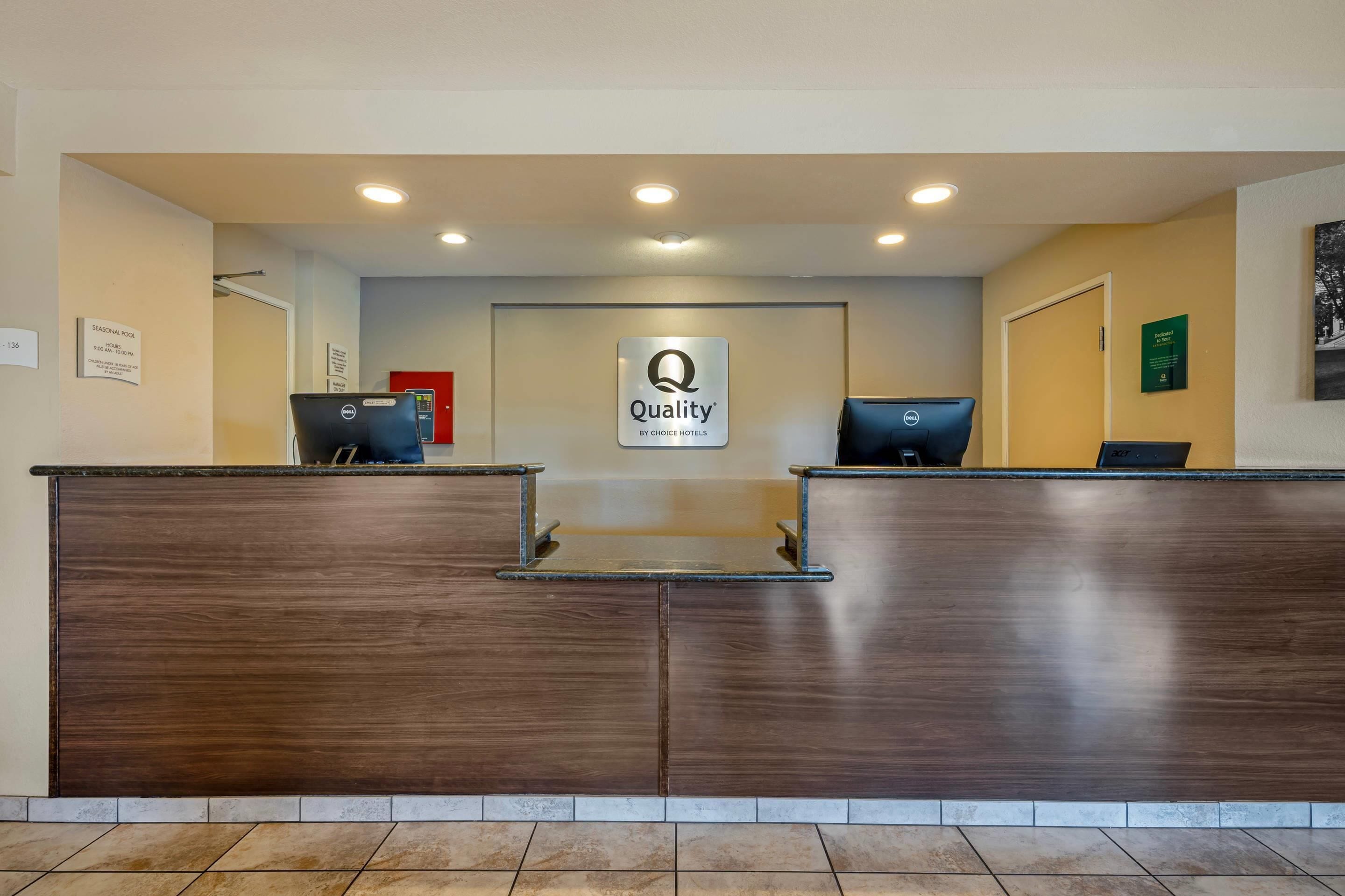 Un mostrador de recepción de hotel con un cartel de "Quality by Choice Hotels", dos computadoras y una zona de recepción con suelo de baldosas.
