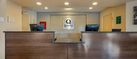 Un mostrador de recepción de hotel con un cartel de "Quality by Choice Hotels", dos computadoras y una zona de recepción con suelo de baldosas.