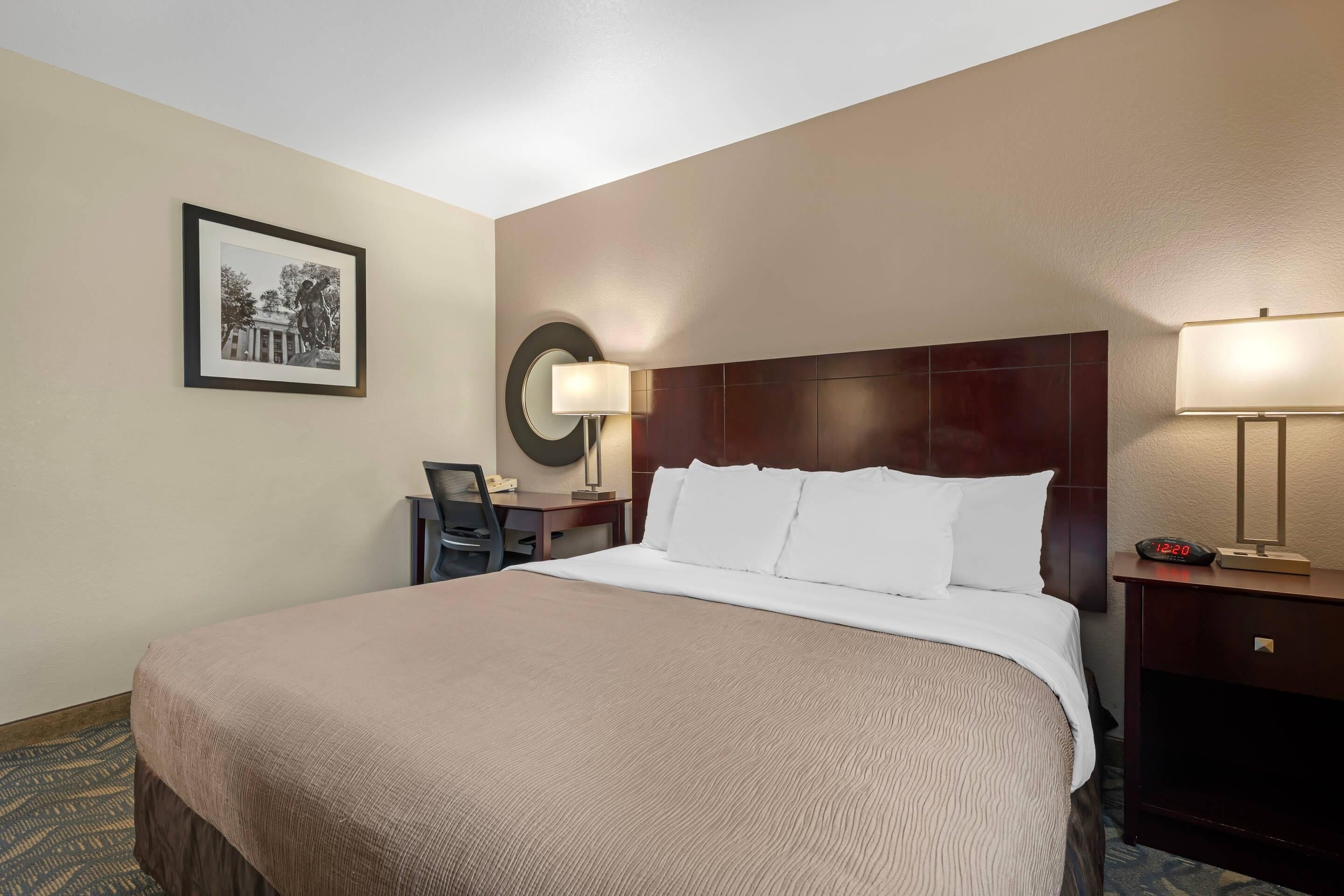 A hotel room with a large bed, a desk with a chair, a lamp, and a framed picture on the wall.