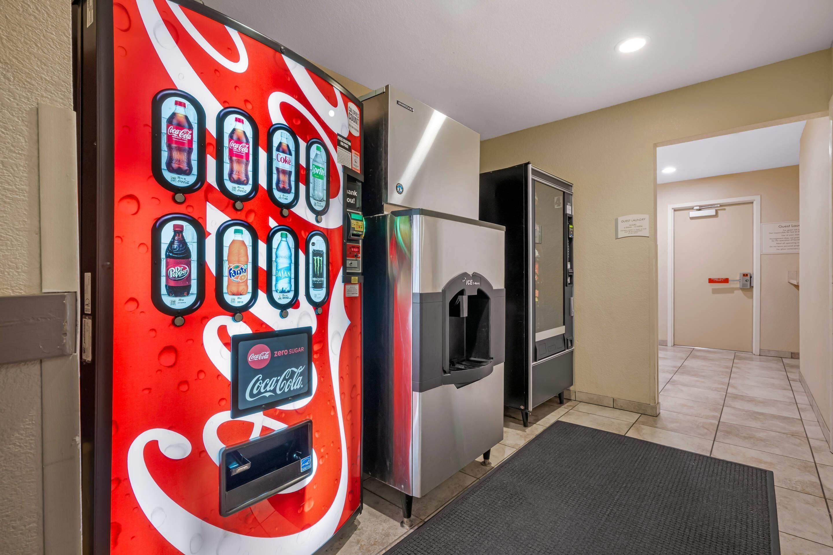 Máquina de vendas com Coca-Cola e outras bebidas, um frigorífico e a porta de um quarto de hóspedes.