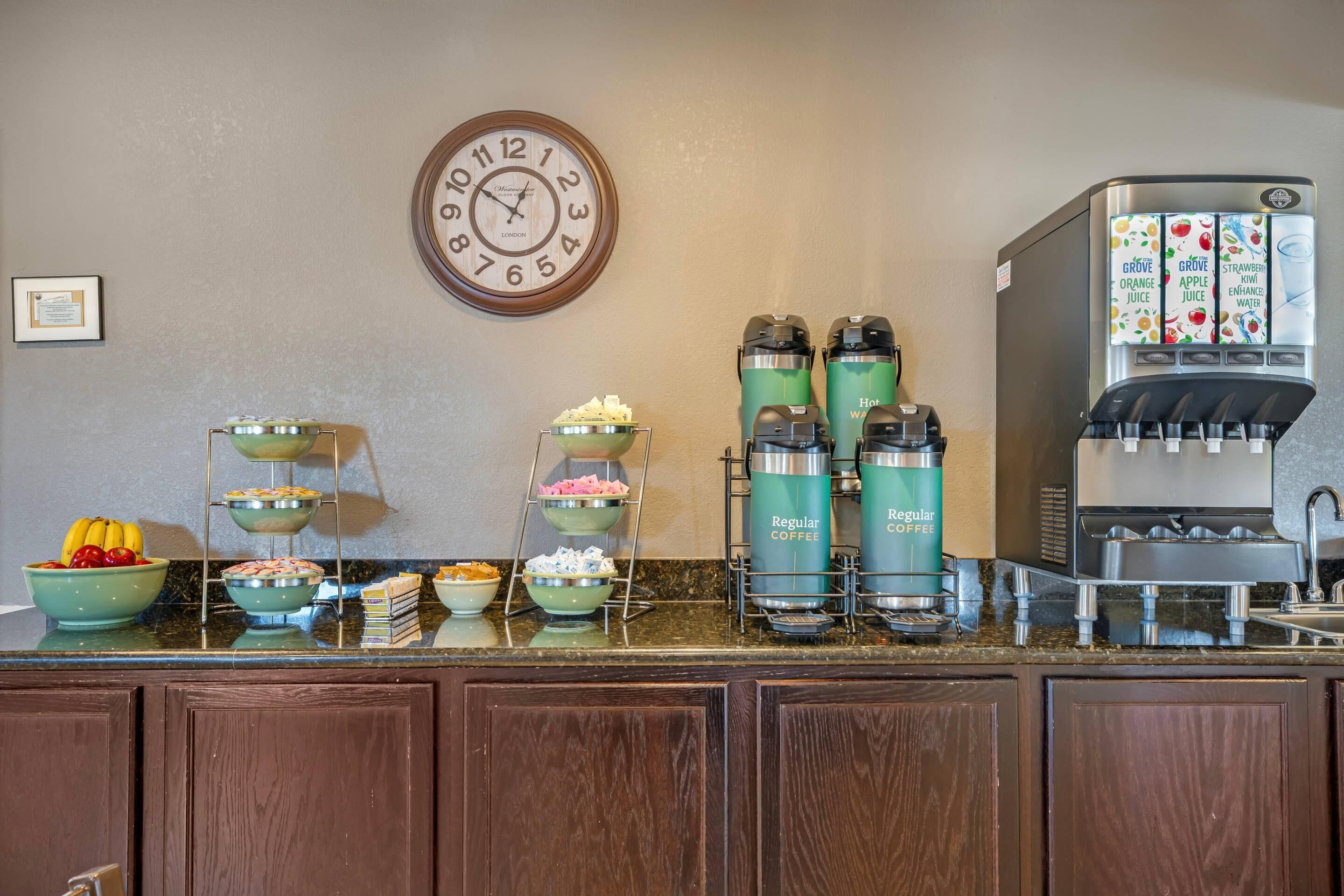 A breakfast bar with coffee makers, fruit bowls, and a clock on the wall.