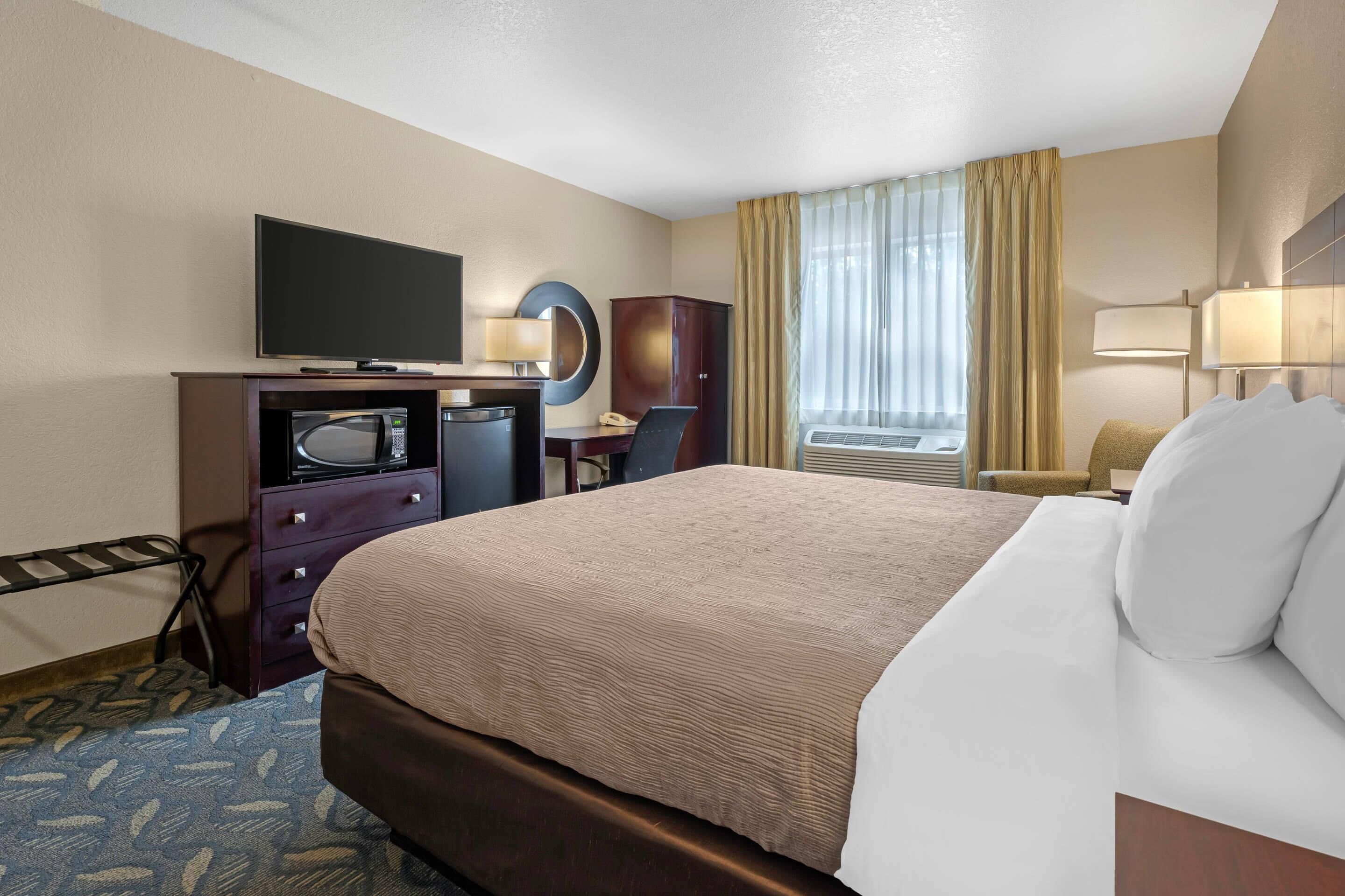 A hotel room with a large bed, a desk with a computer, a television, a minibar, and a window with curtains.