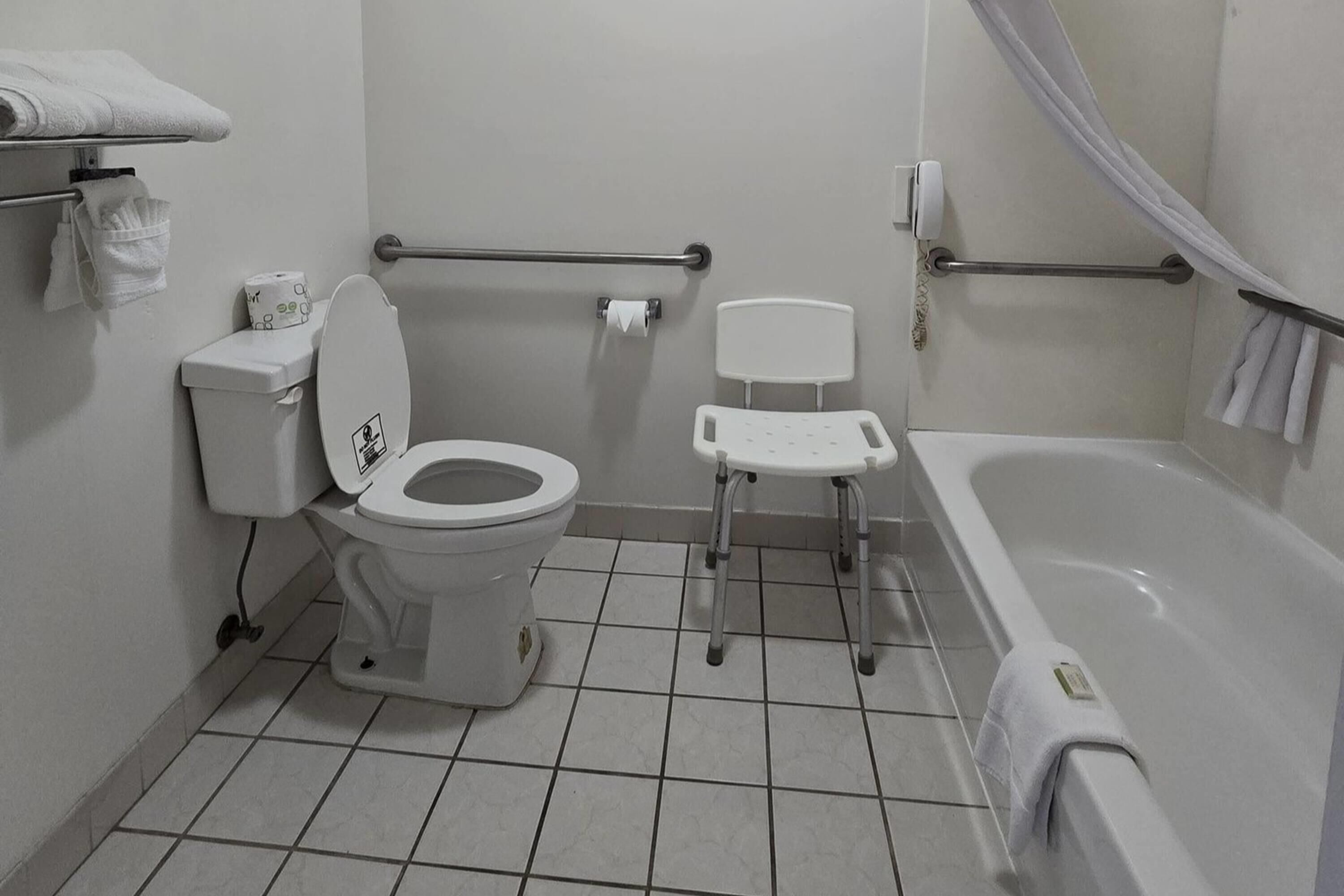 Un baño con inodoro, asiento de inodoro elevado, barra de apoyo, bañera y cortina de ducha.