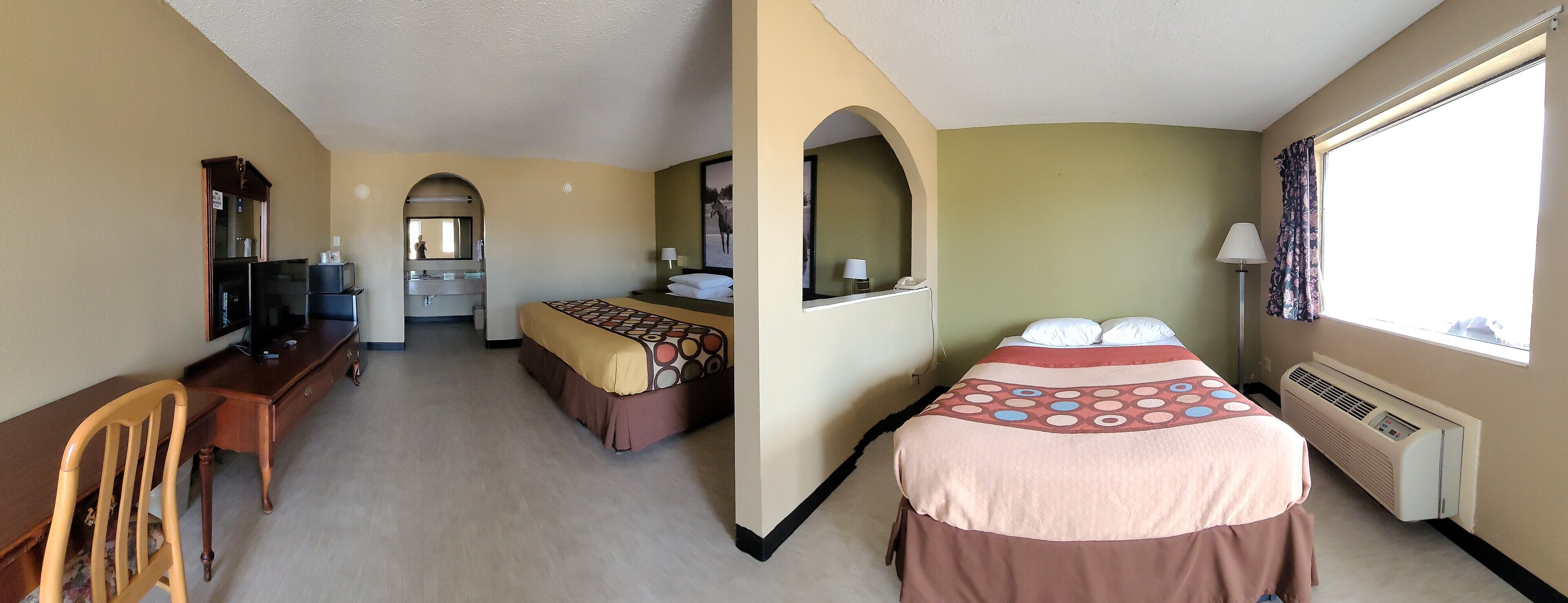 A hotel room with a bed, a desk, a chair, a mirror, a window, and a wall-mounted heater.