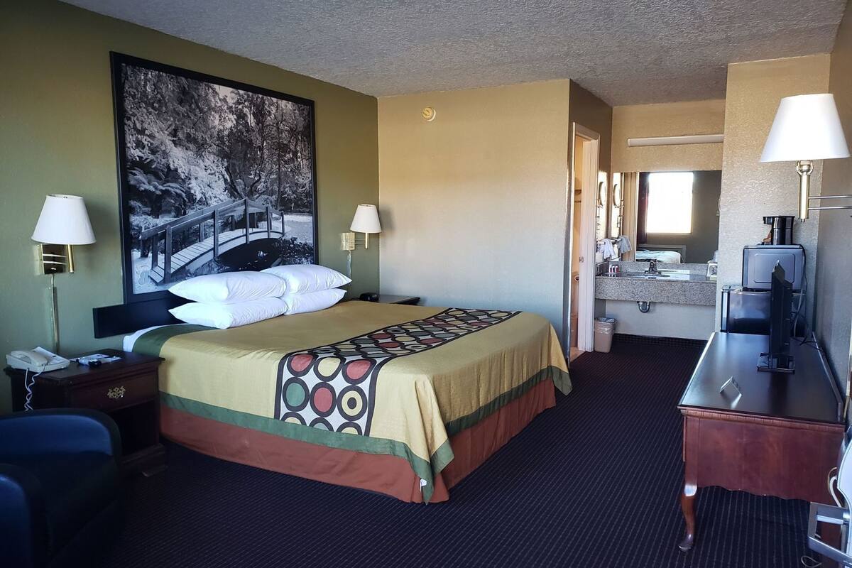 Une chambre d’hôtel avec un lit, deux lampes de chevet, un bureau avec une lampe, un miroir et une salle de bain visible à travers une porte ouverte.