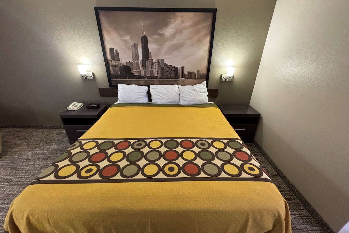 A hotel room with a bed, two bedside tables, a cityscape painting, and two wall-mounted lights.