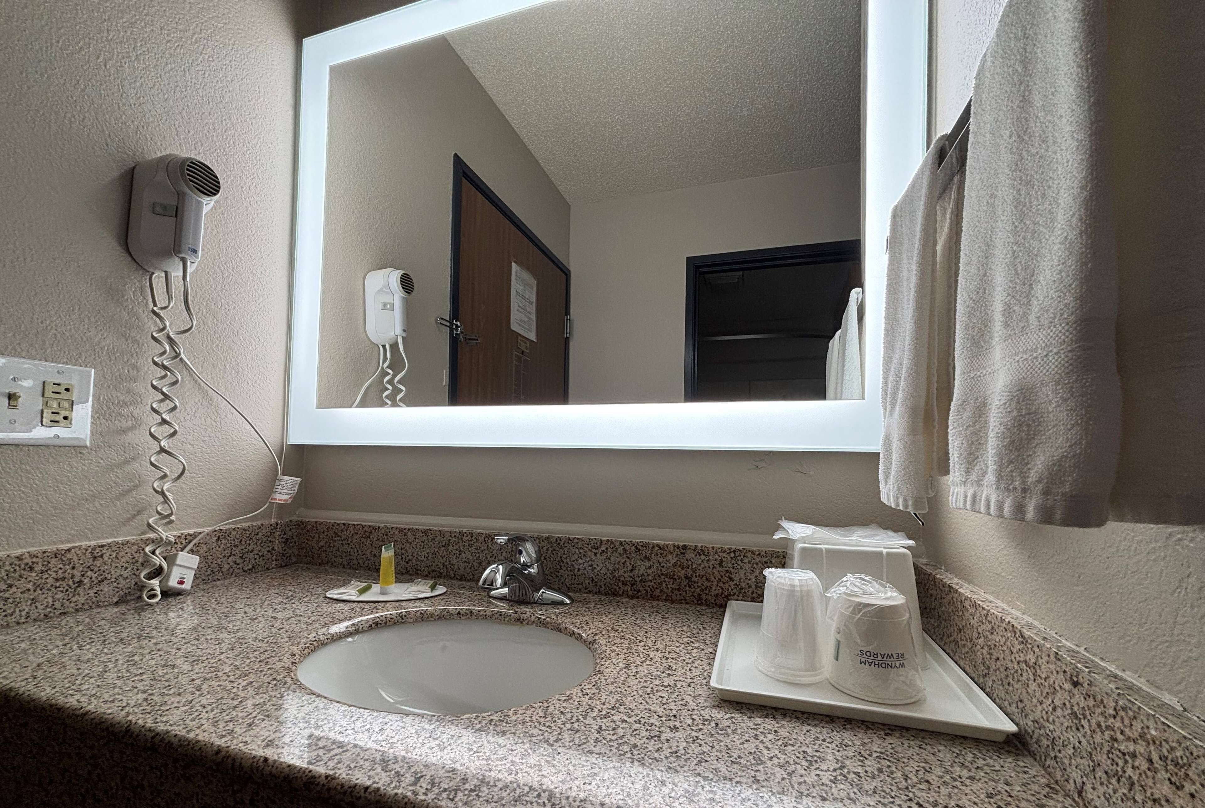 Une salle de bain d’hôtel avec un comptoir en granit, un lavabo, un miroir, un sèche-cheveux et des articles de toilette disposés sur un plateau.
