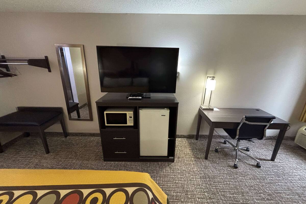 A hotel room with a flat-screen TV, a small refrigerator, a desk, a chair, a bed, and a mirror.