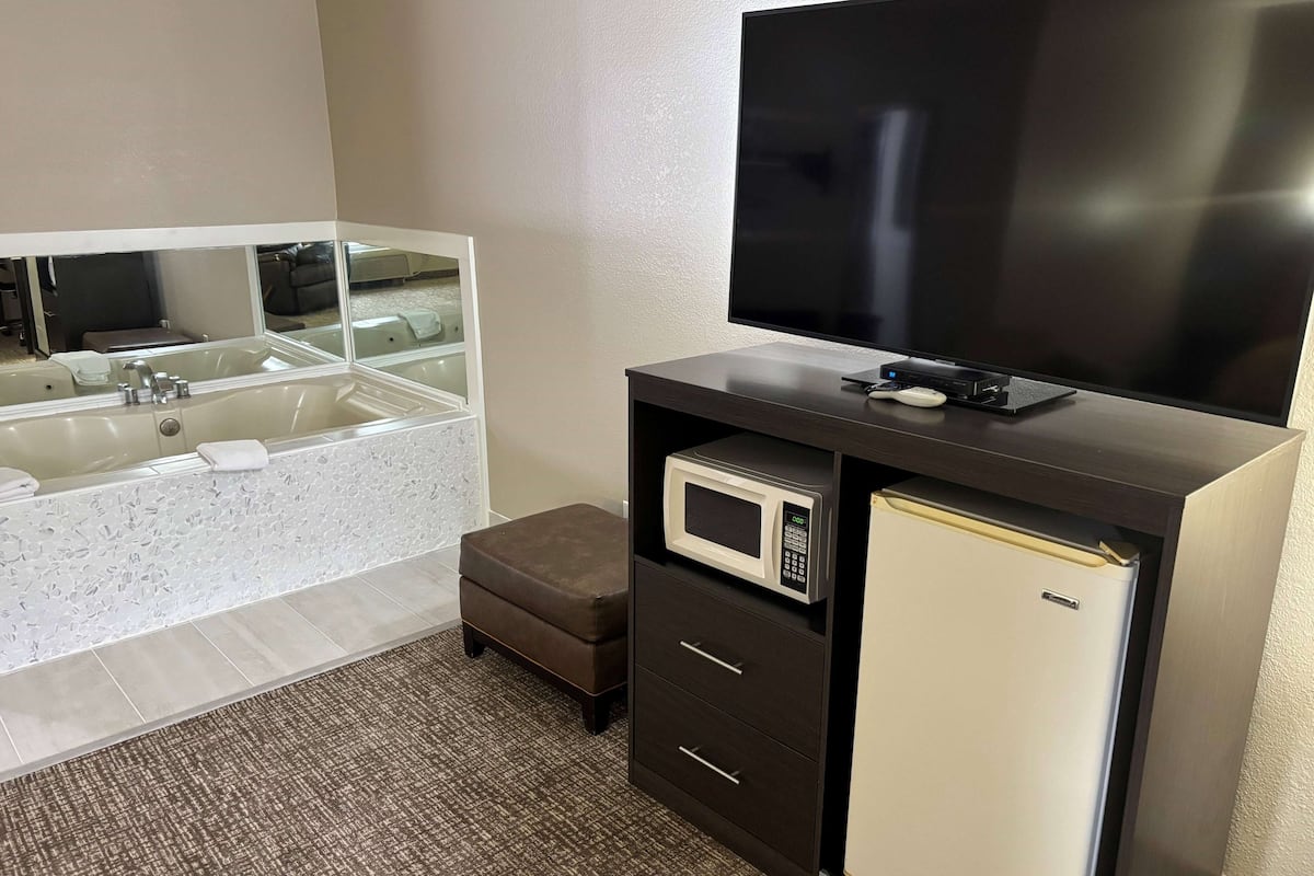 A bathroom with a large mirror, a bathtub, a toilet, a microwave, a refrigerator, and a flat-screen TV.