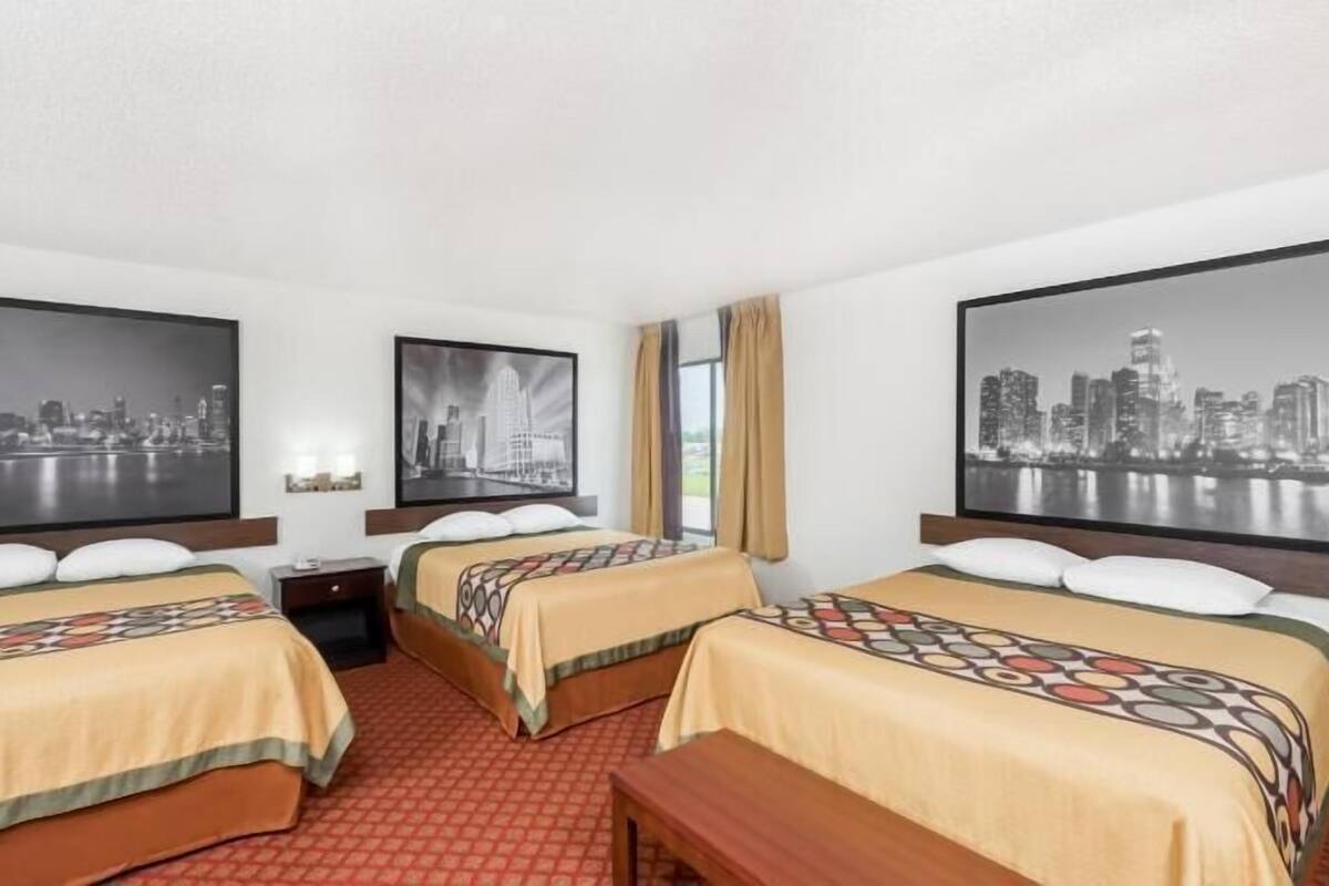 A hotel room with three beds, each with a patterned bedspread, a wooden bench, and a nightstand. There are three large framed cityscape photographs on the walls.