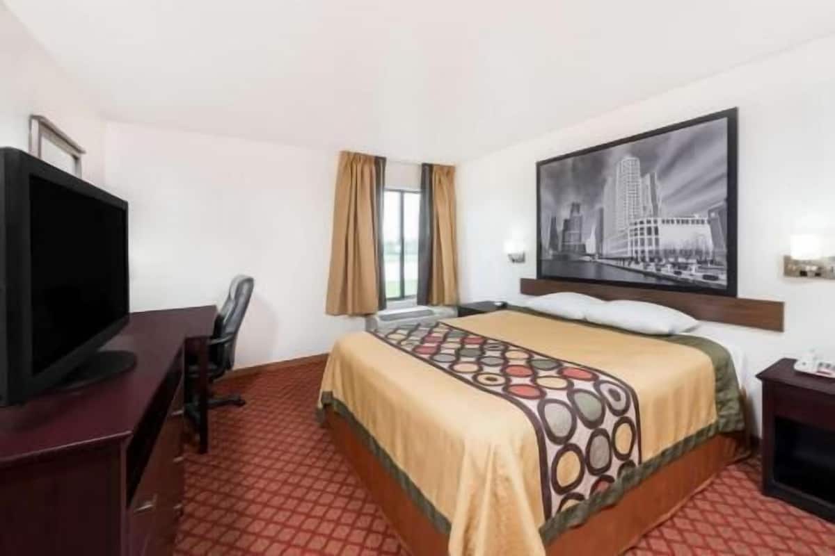 A hotel room with a bed, a desk with a TV, a chair, a window with curtains, and a cityscape painting on the wall.