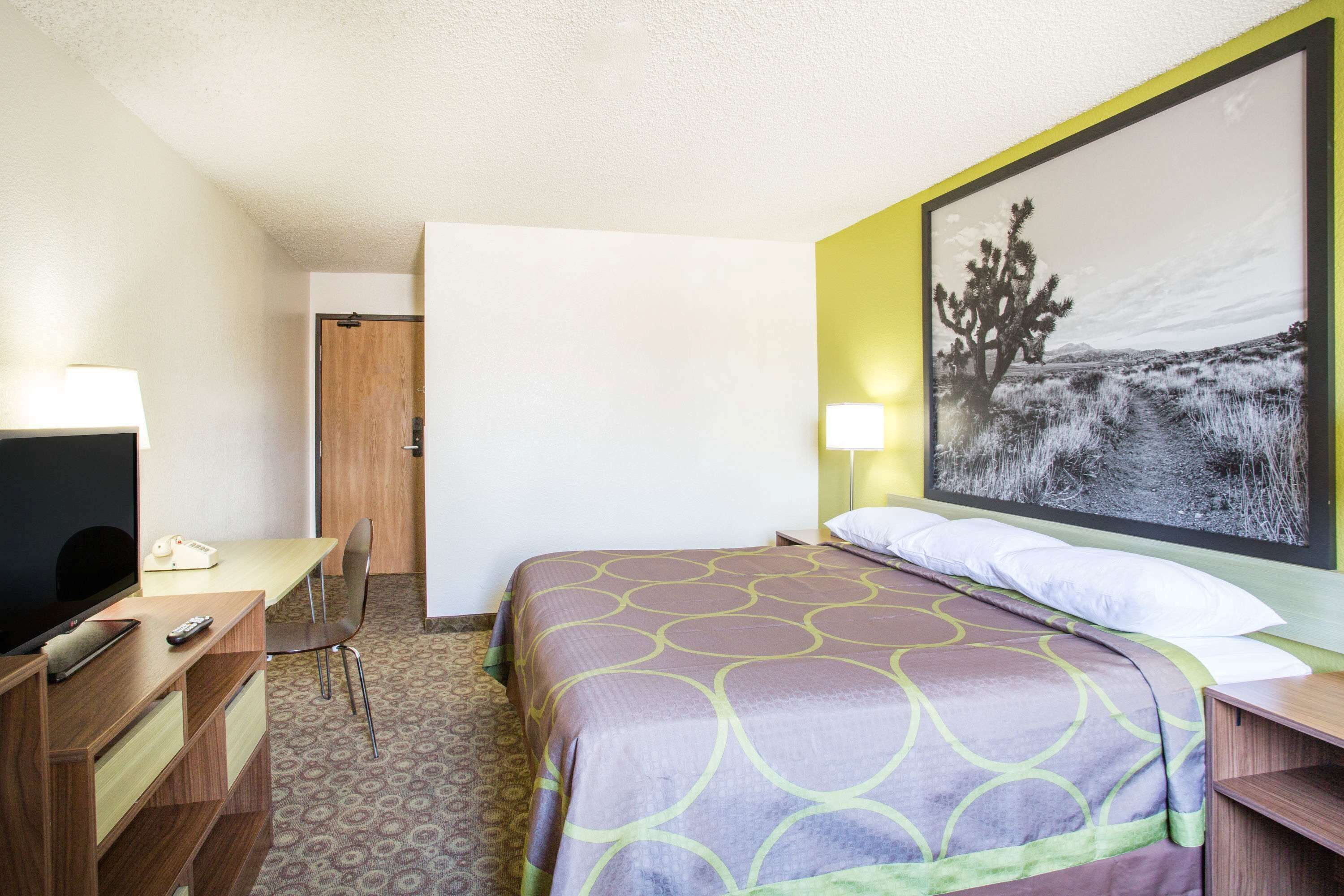 A hotel room with a bed, a desk, a chair, a television, and a large framed picture on the wall.