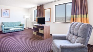 Habitación de hotel con sofá, una silla, televisor sobre una cómoda de madera, lámpara y ventanas amplias con cortinas.