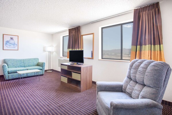 A hotel room with a sofa, a chair, a TV on a wooden cabinet, a lamp, and large windows with curtains.