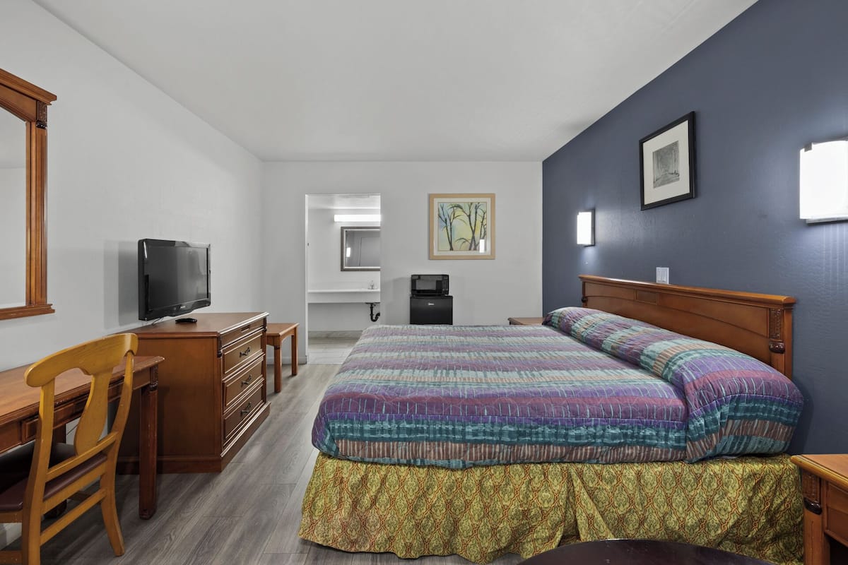 A hotel room with a bed, a desk with a television, a chair, a dresser, a microwave, and a painting on the wall.