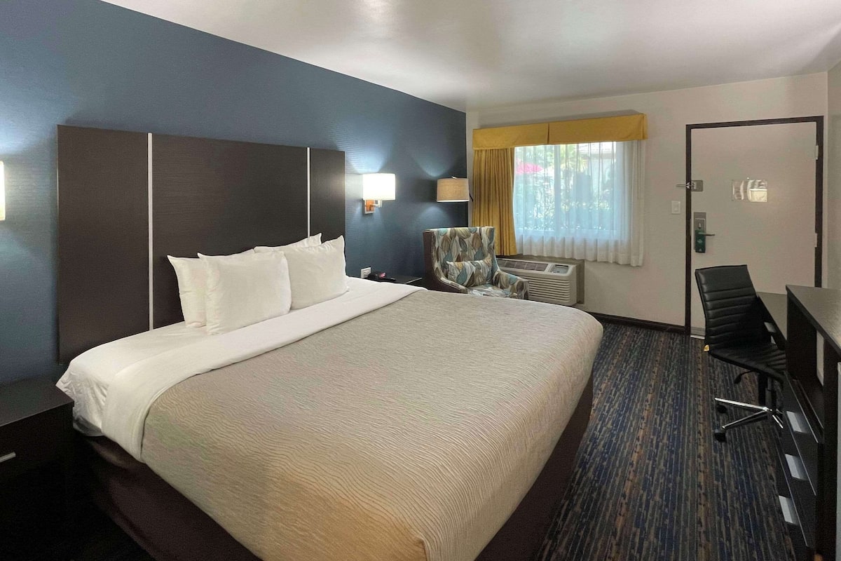 A hotel room with a large bed, a desk, a chair, a lamp, and a window with curtains.