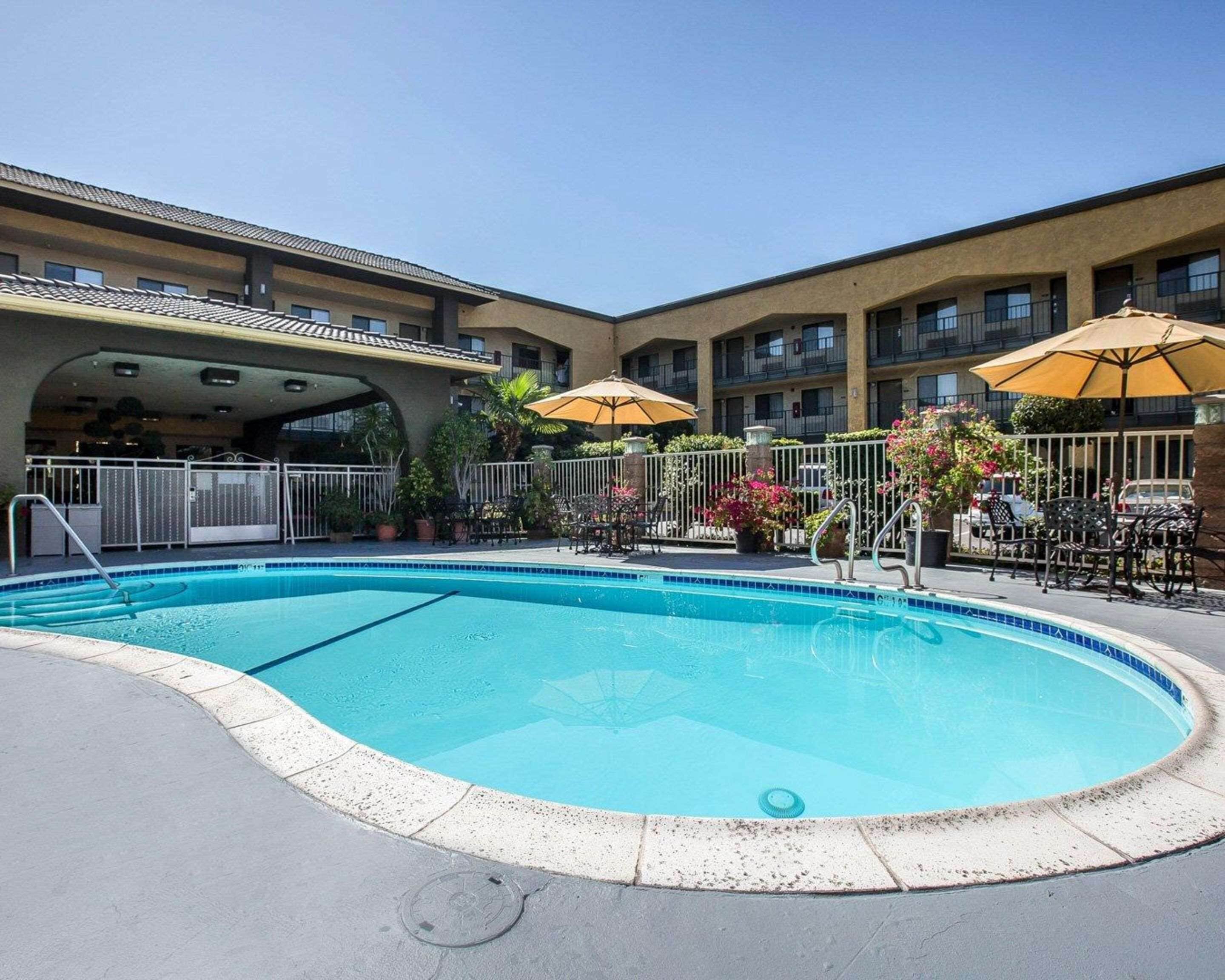 A pool area with a clear blue pool, surrounded by outdoor seating and umbrellas, and a building with multiple balconies in the background.
