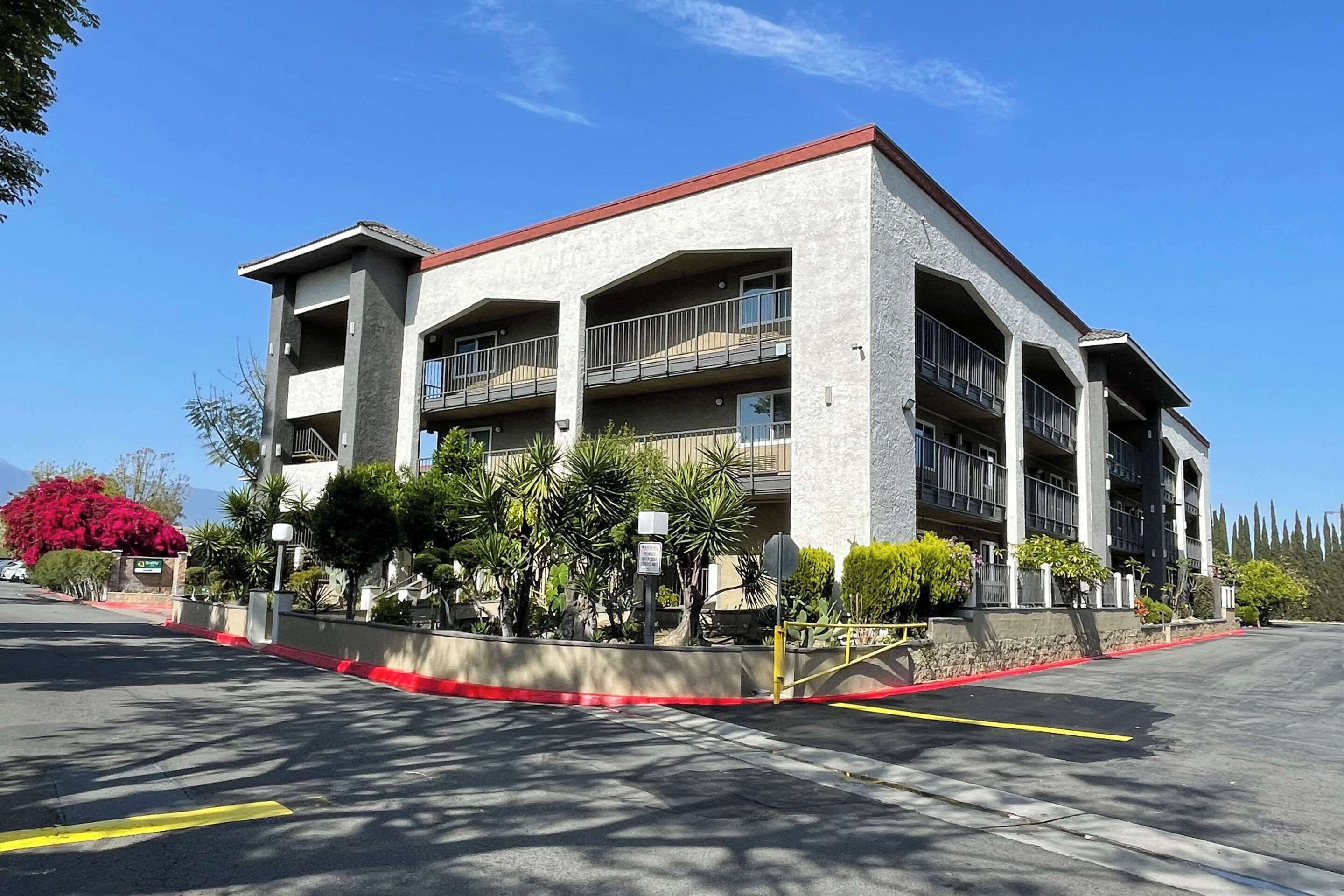 A multi-story hotel with balconies, a parking lot, and landscaped areas with various plants.