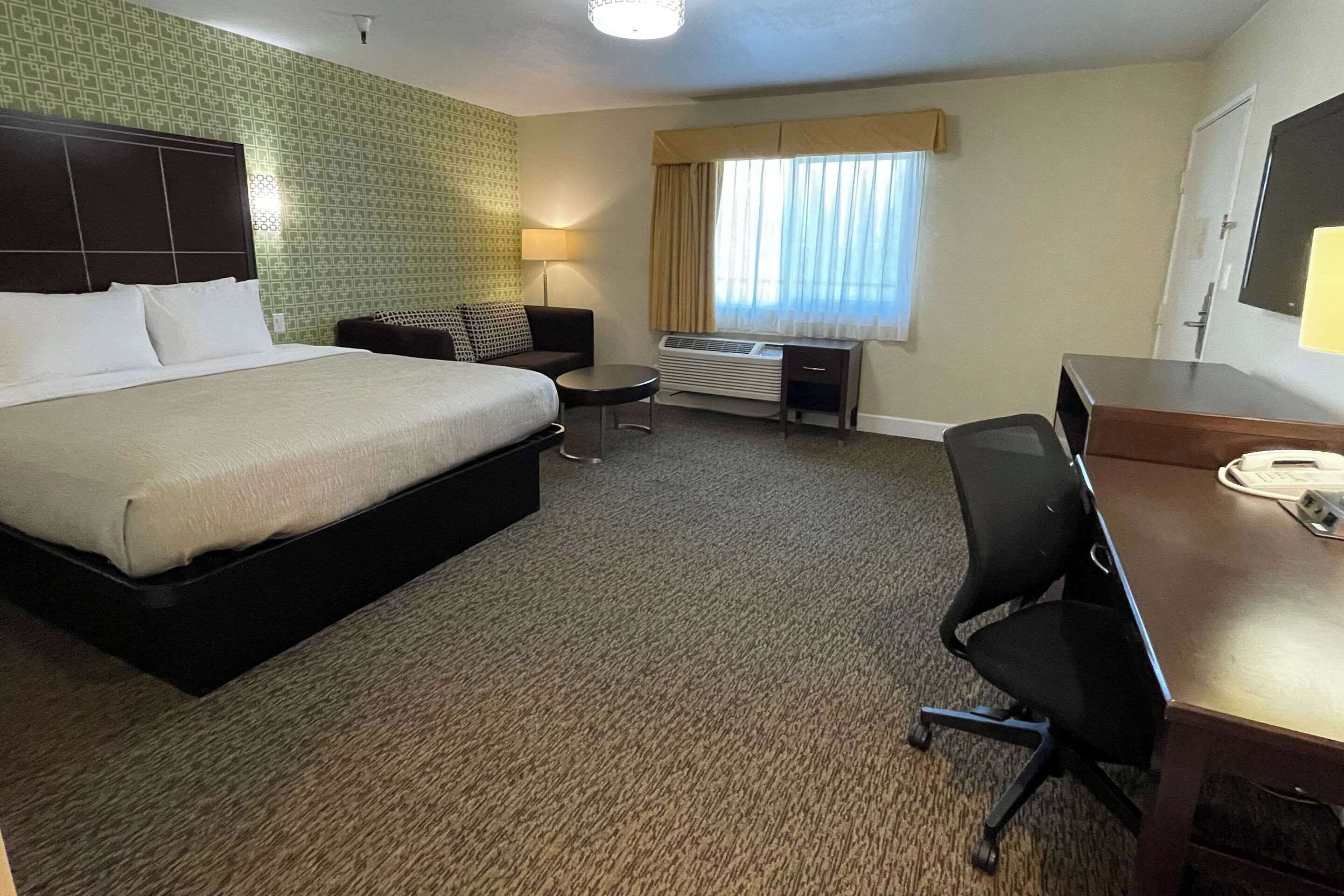 A hotel room with a large bed, a desk with a telephone, a chair, a small table, a lamp, and a window with curtains.