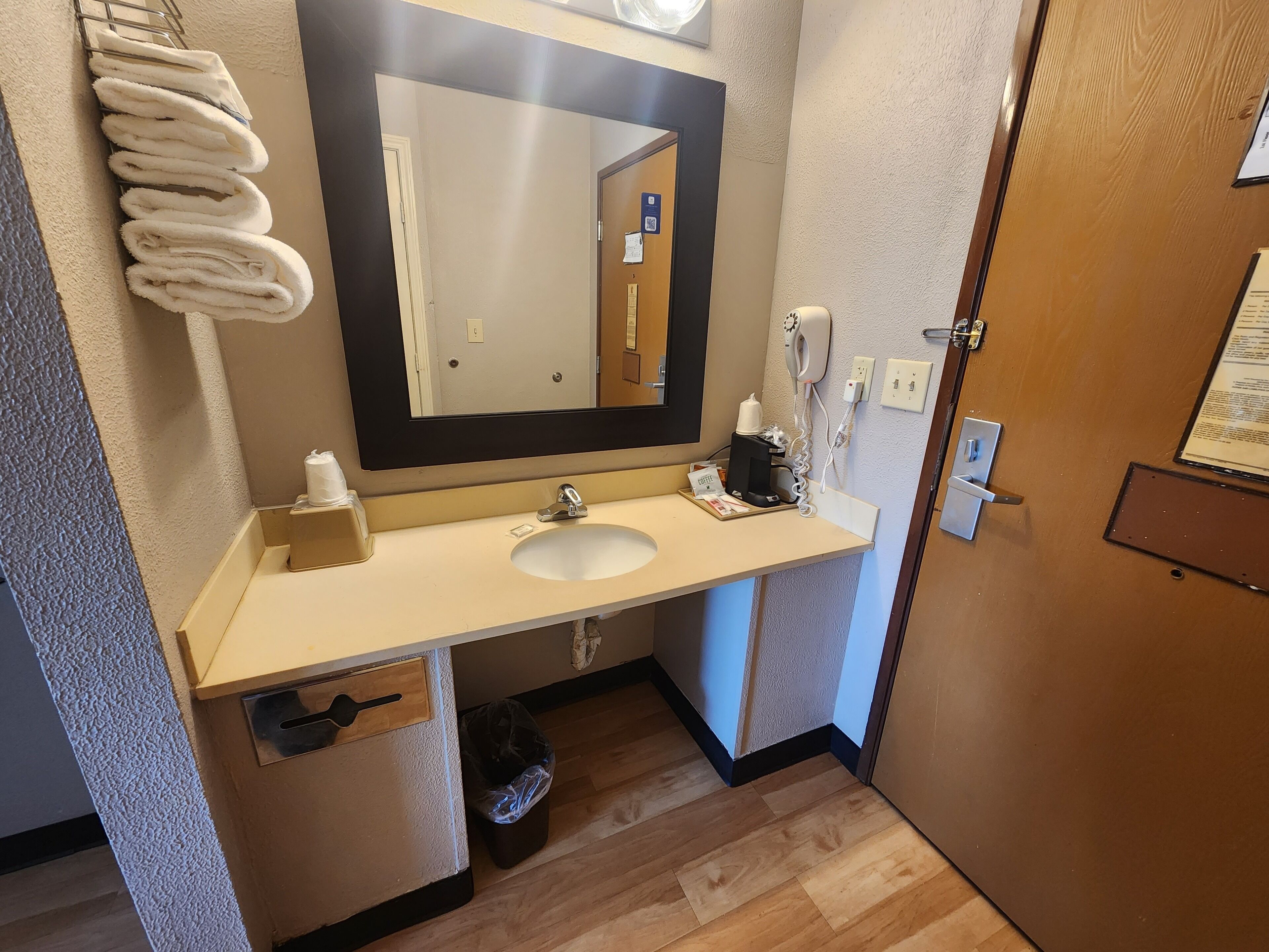 Un baño de hotel con lavamanos, espejo, toallero y un cubo de basura.