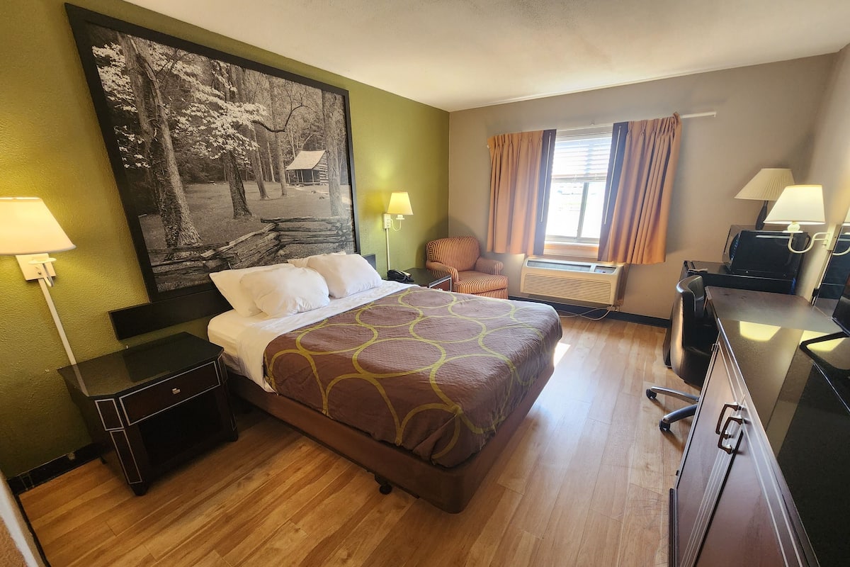 A hotel room with a large bed, a desk, a chair, a desk lamp, a wall-mounted air conditioner, and a window with curtains.