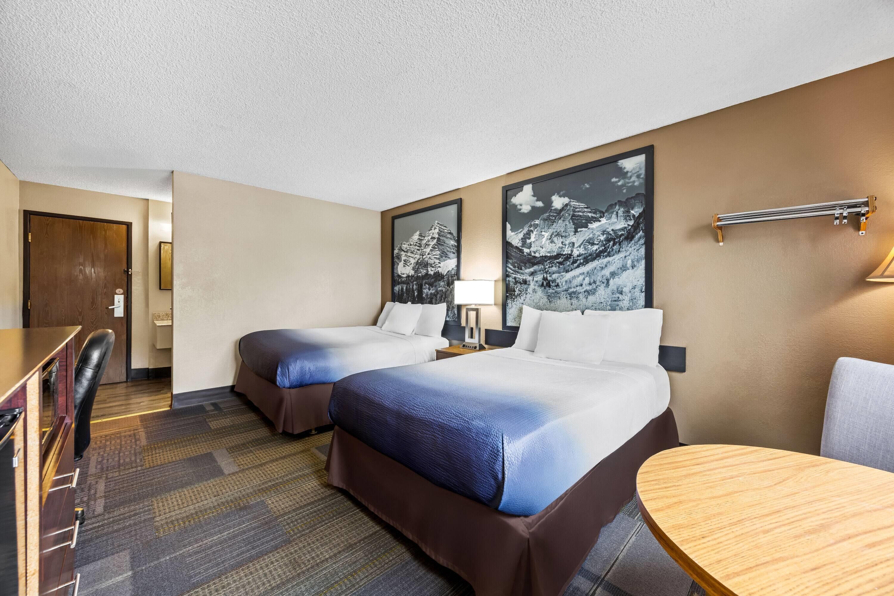 Quarto de hotel com duas camas, uma escrivaninha e uma cadeira. Há duas molduras com imagens na parede.