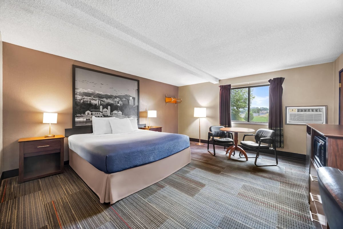 Ein Hotelzimmer mit einem großen Bett, zwei Nachttischen mit Lampen, einem Schreibtisch mit zwei Stühlen, einem Fenster mit Vorhängen und einem gerahmten Bild an der Wand.