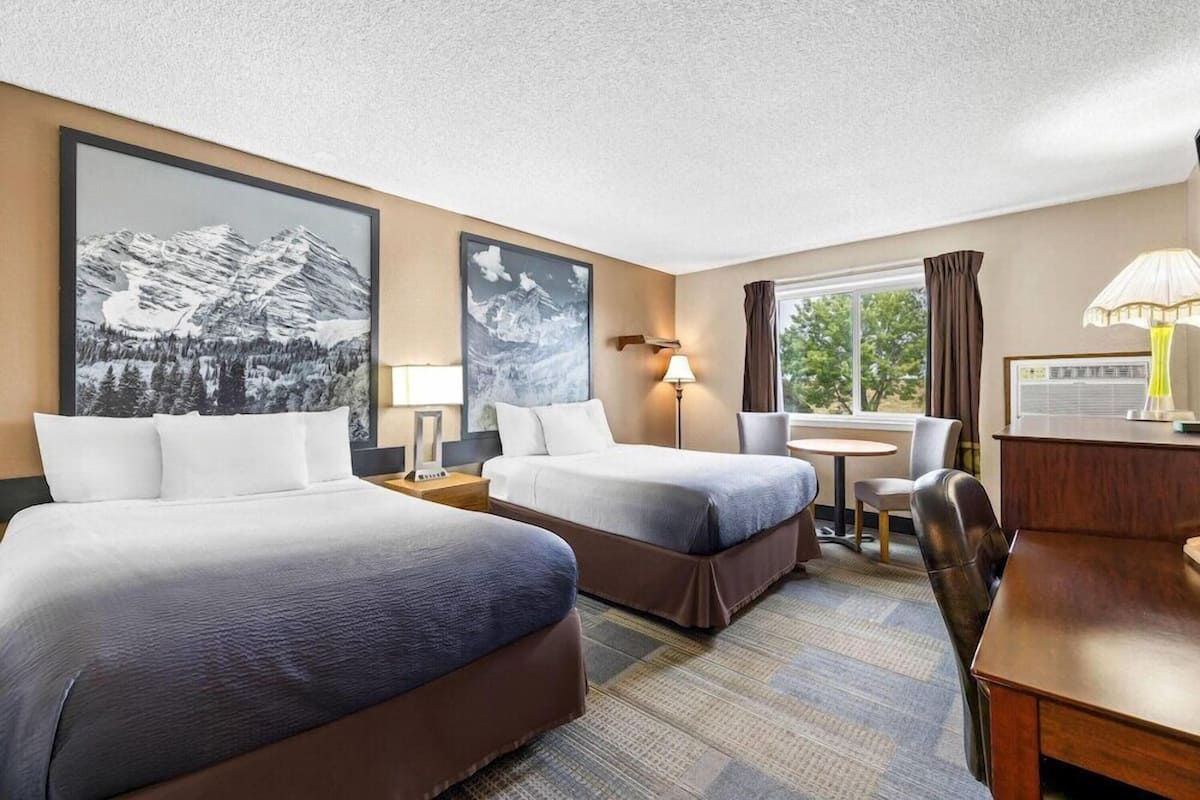 Ein Hotelzimmer mit zwei Betten, einem Schreibtisch, einem Stuhl, einer Lampe und einem Fenster mit Vorhängen.