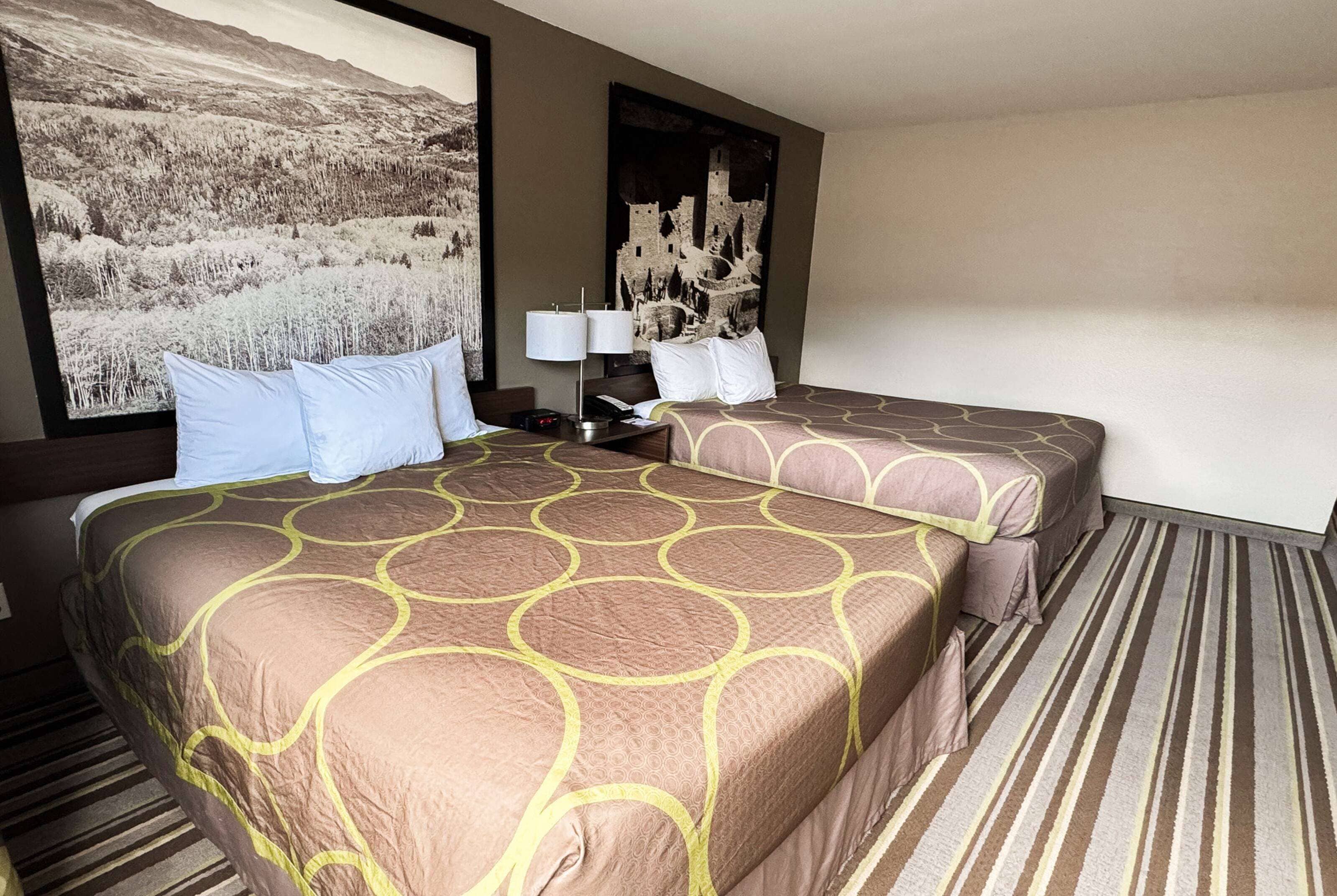 Una camera d'albergo con due letti, un'ampia finestra vista montagna, un comodino con una lampada e un'opera d'arte appesa al muro.