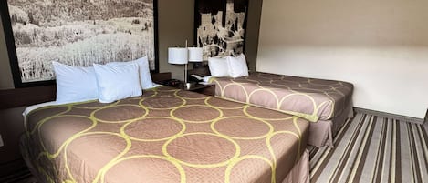 Una camera d'albergo con due letti, un'ampia finestra vista montagna, un comodino con una lampada e un'opera d'arte appesa al muro.