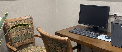Un escritorio de madera con una computadora, que incluye un monitor, teclado y ratón. Hay dos sillas con estampado, con estructura de madera y asientos acolchados.