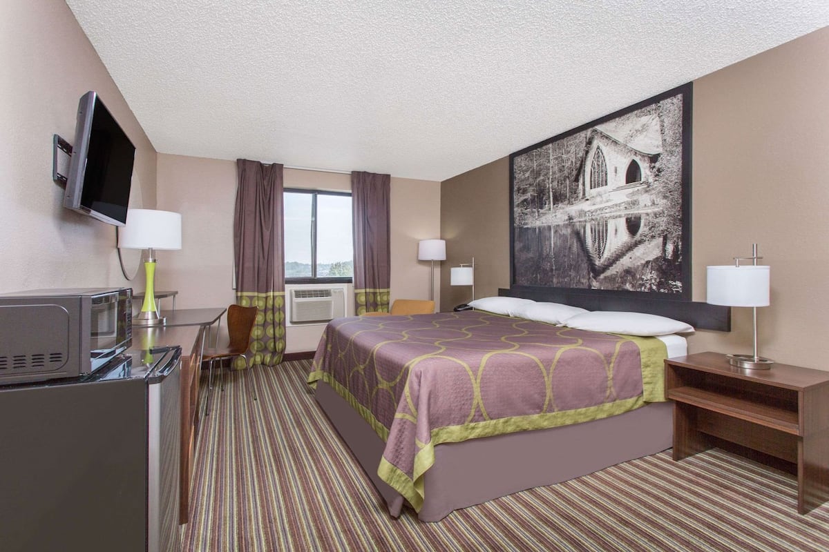 Una habitación de hotel con una cama, un escritorio con televisor, una ventana con cortinas y un cuadro grande en la pared.