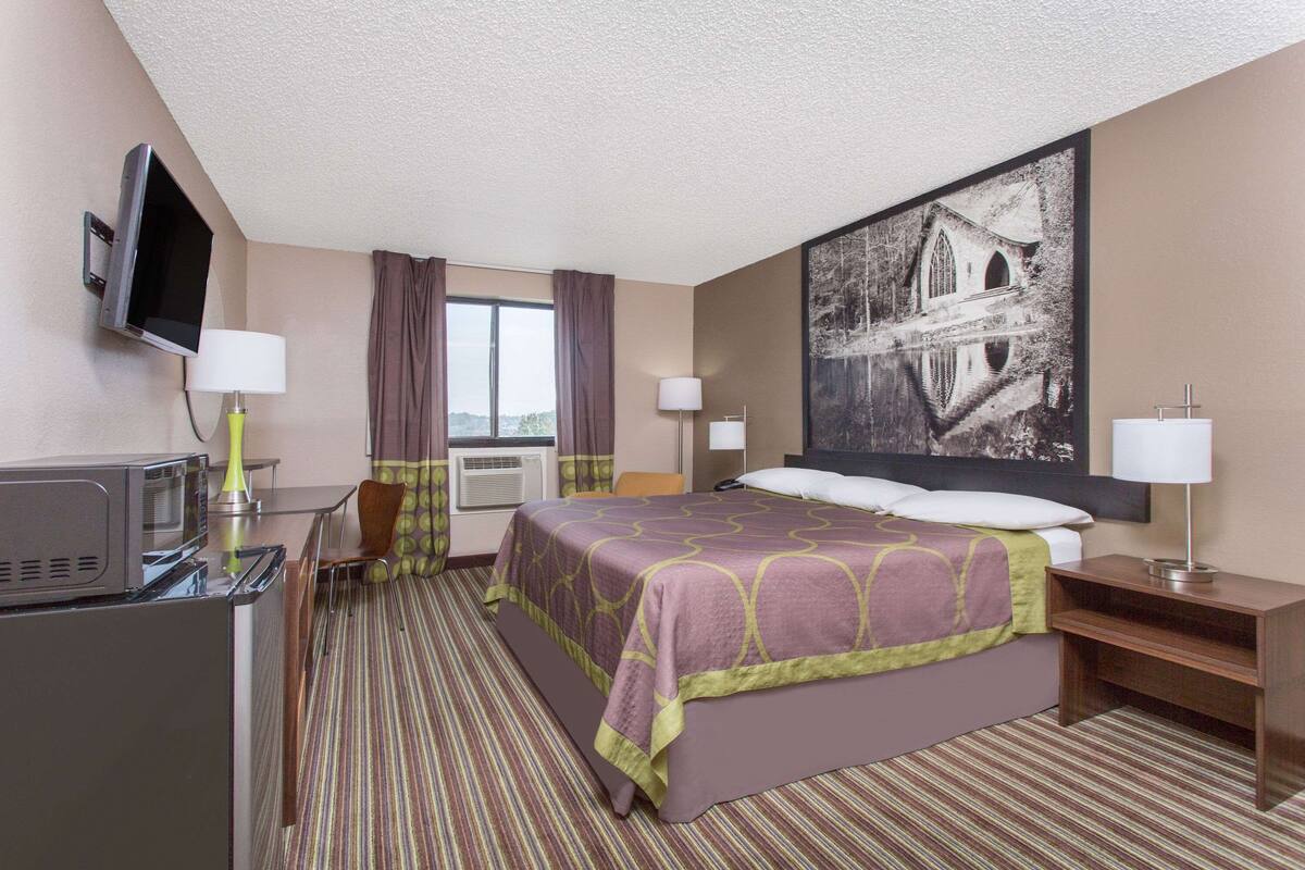 Una habitación de hotel con cama, escritorio con televisor, silla, ventana con cortinas y un cuadro en la pared.