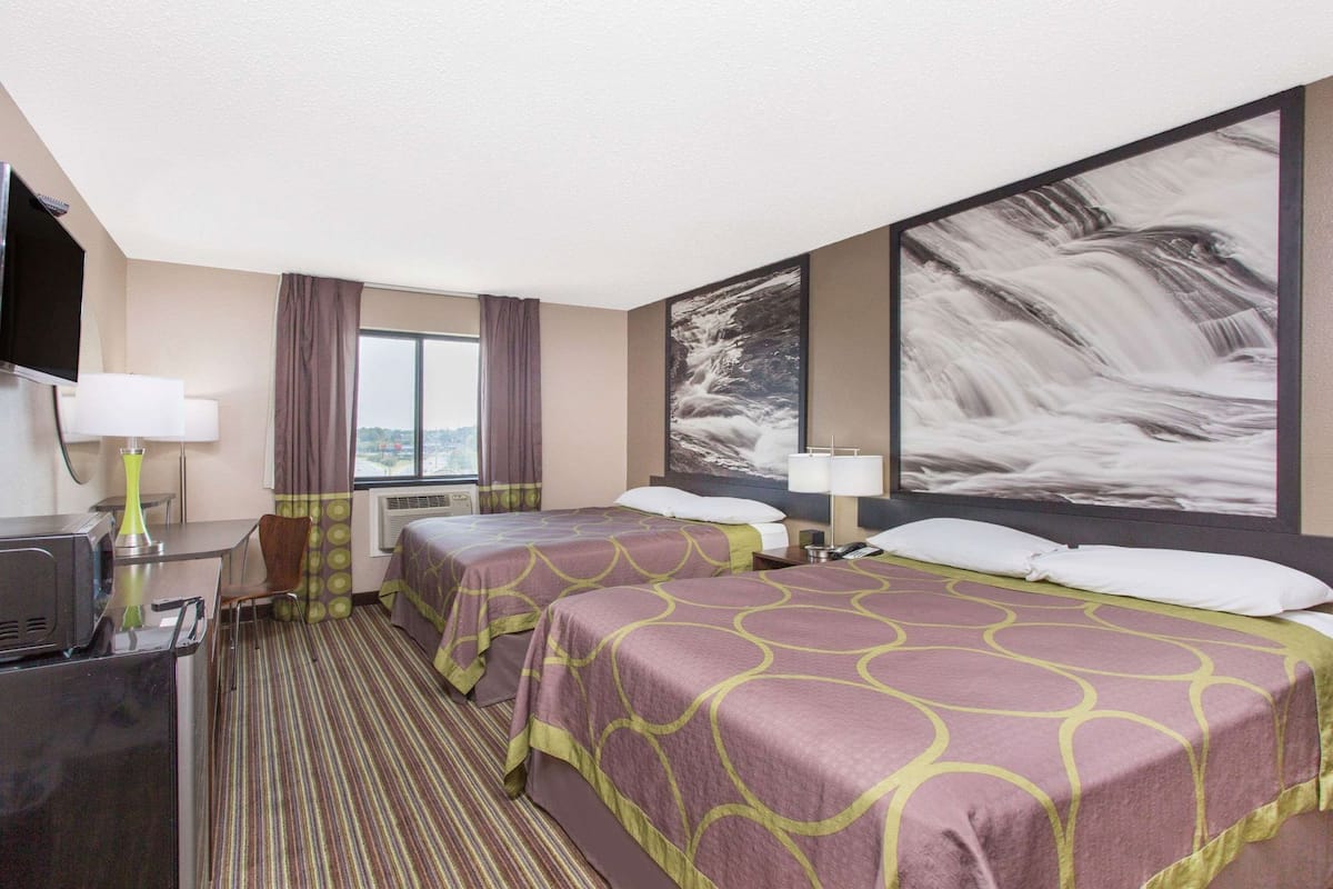 Habitación de hotel con dos camas, un escritorio, una silla, un televisor y un ventanal grande con cortinas.