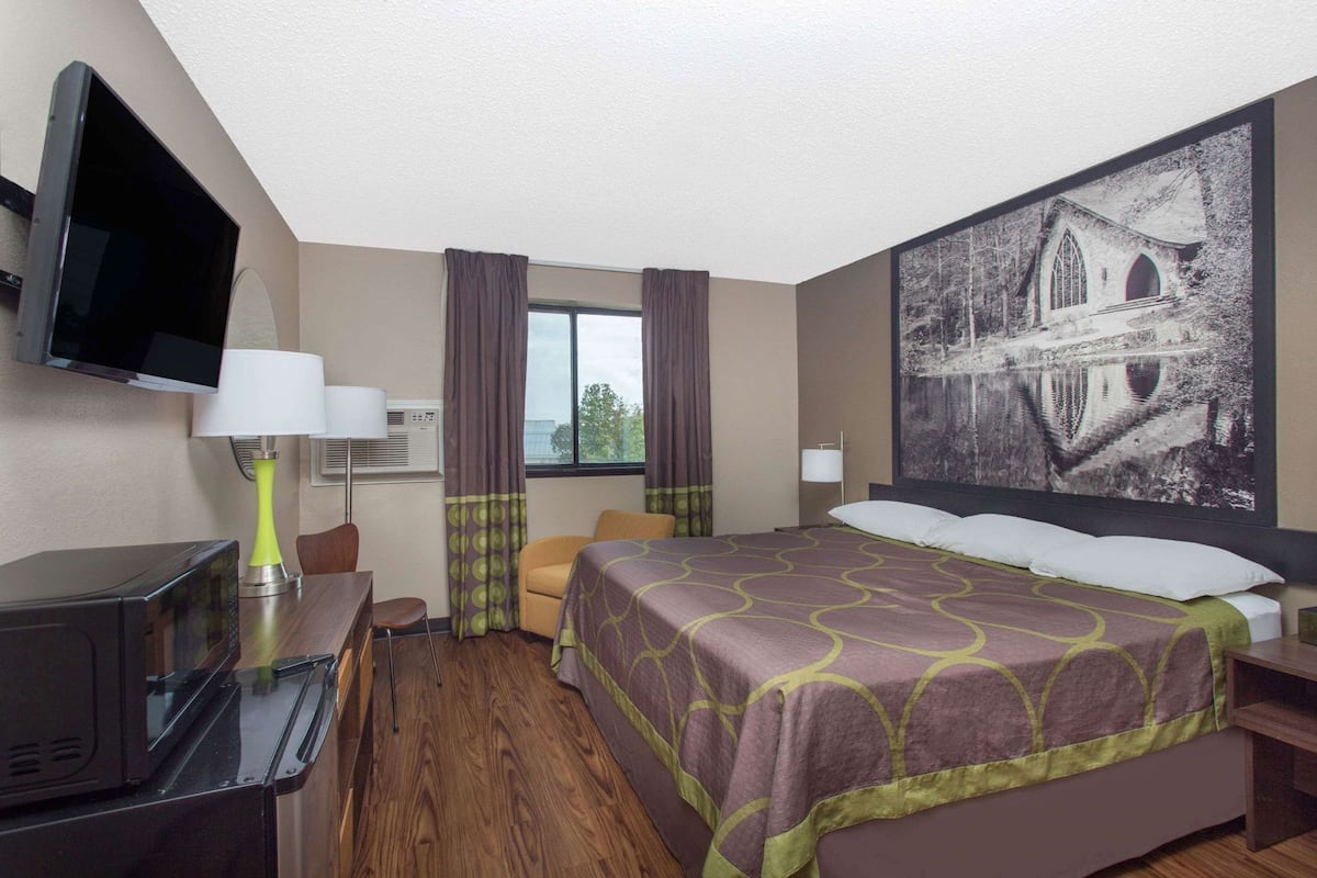Habitación de hotel con cama, televisión, escritorio con lámpara, silla, ventana con cortinas y un cuadro grande en la pared.