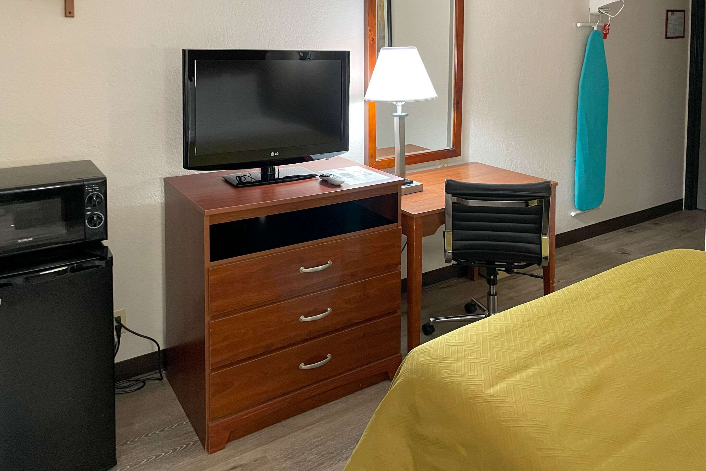 Habitación de hotel con escritorio de madera, televisor, lámpara, refrigerador pequeño, silla y cama con cubierta amarilla.