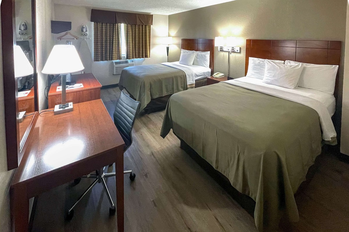 Habitación de hotel con dos camas, un escritorio, una silla, una lámpara, un televisor de pantalla plana y una ventana con cortinas.