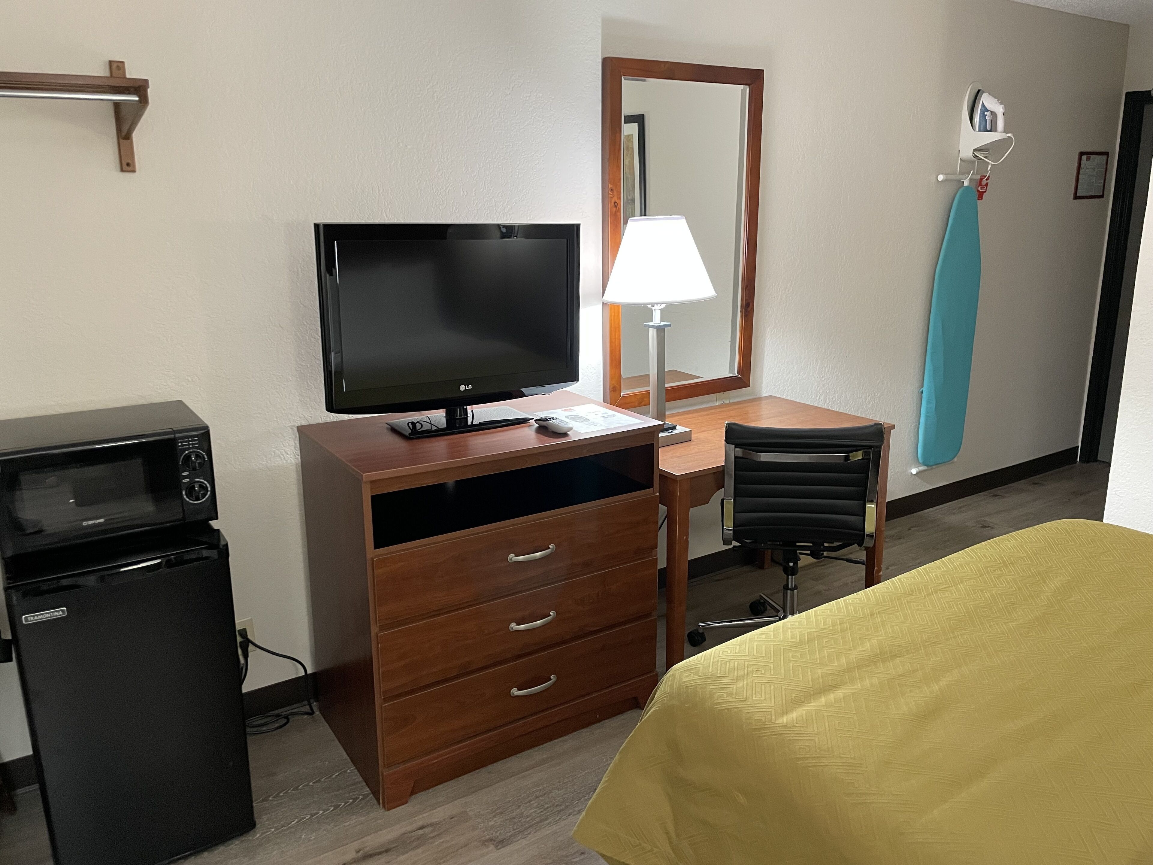 A hotel room with a wooden desk, a television, a lamp, a microwave, a small refrigerator, a chair, and a bed with a yellow cover.