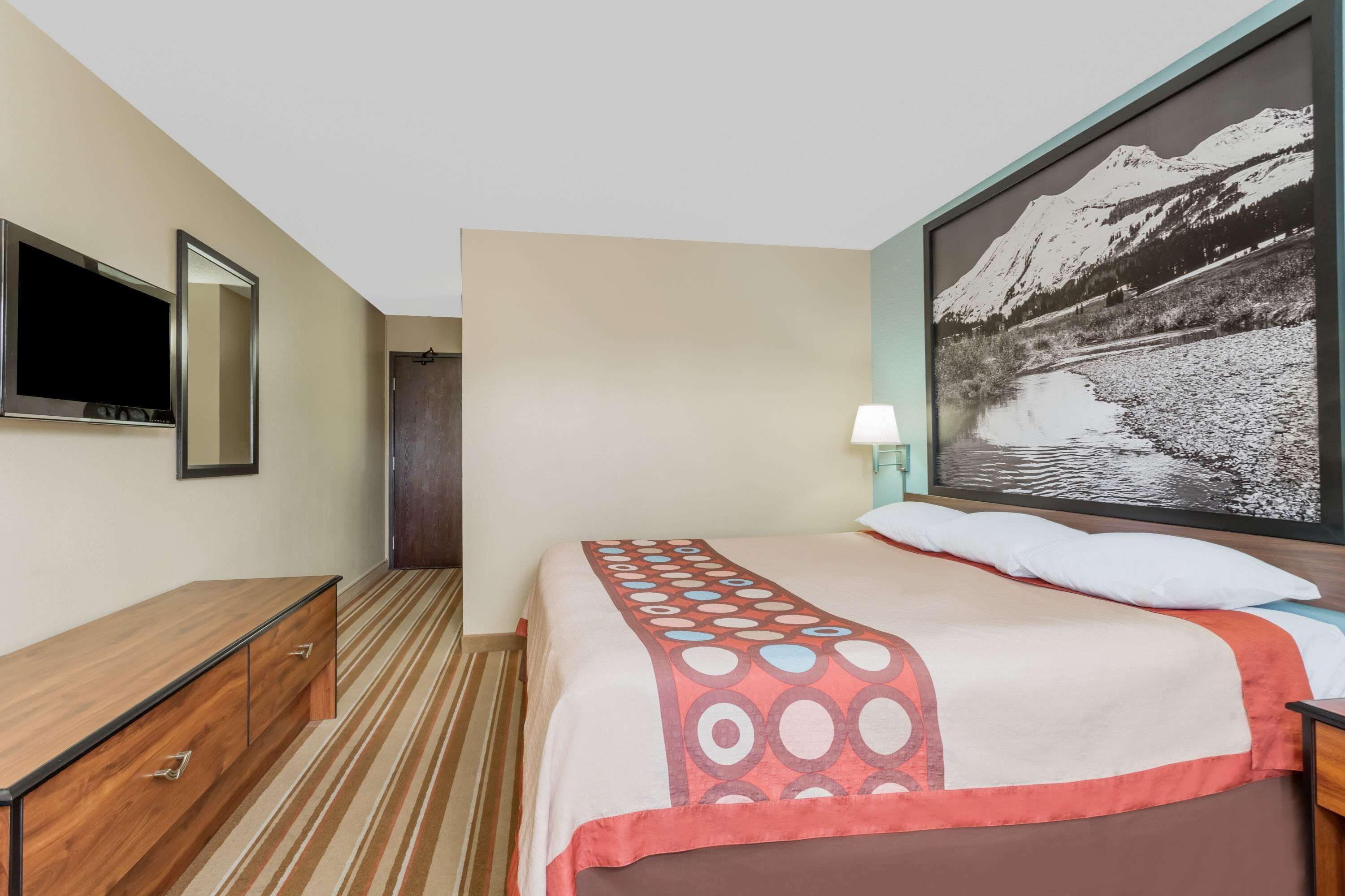 Una habitación de hotel con una cama, un televisor, un aparador de madera, un cuadro de un paisaje montañoso y una lámpara.