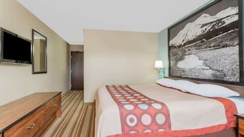 A hotel room with a bed, a TV, a wooden dresser, a painting of a mountain landscape, and a lamp.